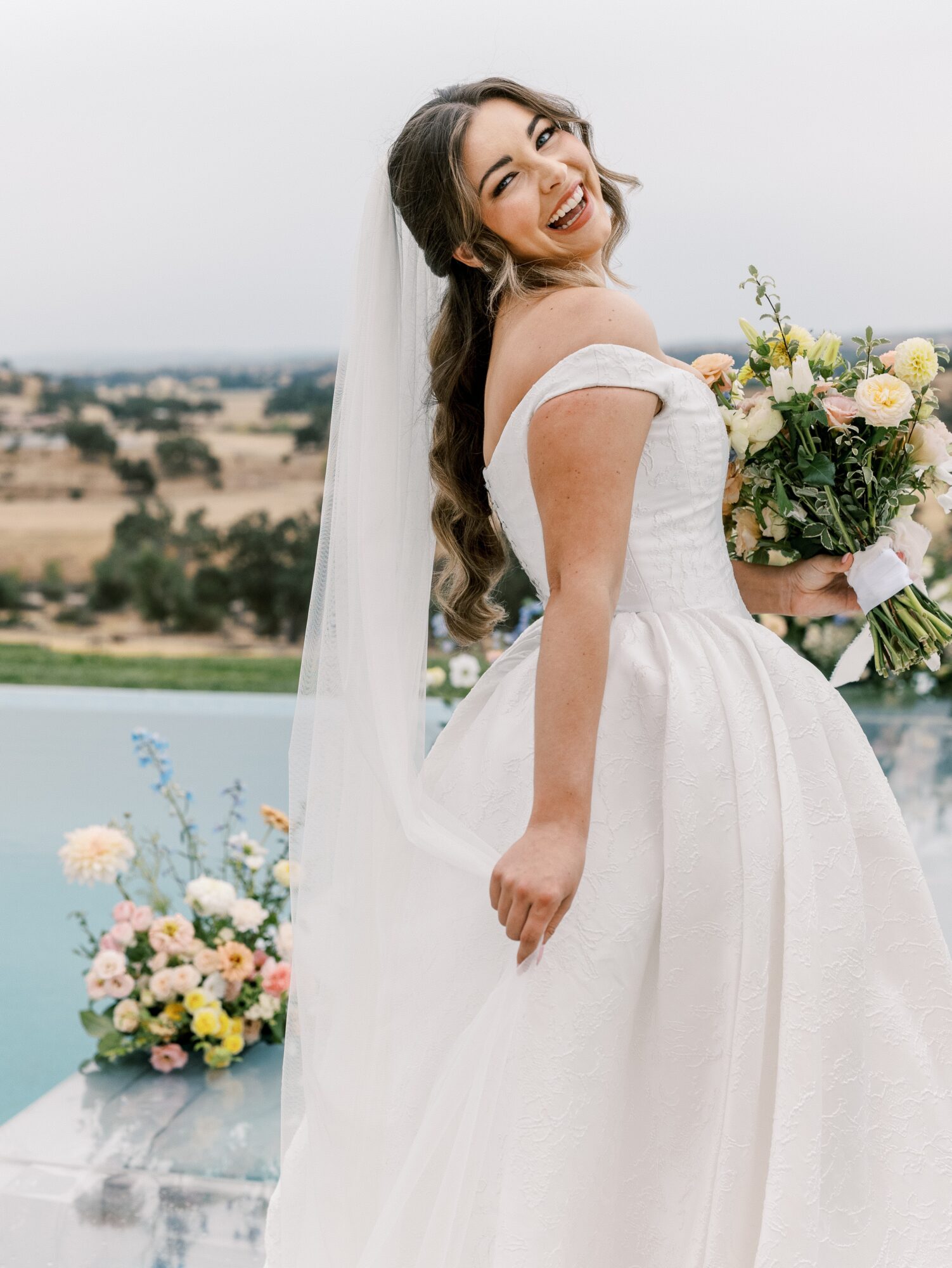 Smiling bride in wedding dress holding bouquet outdoors with landscape background.