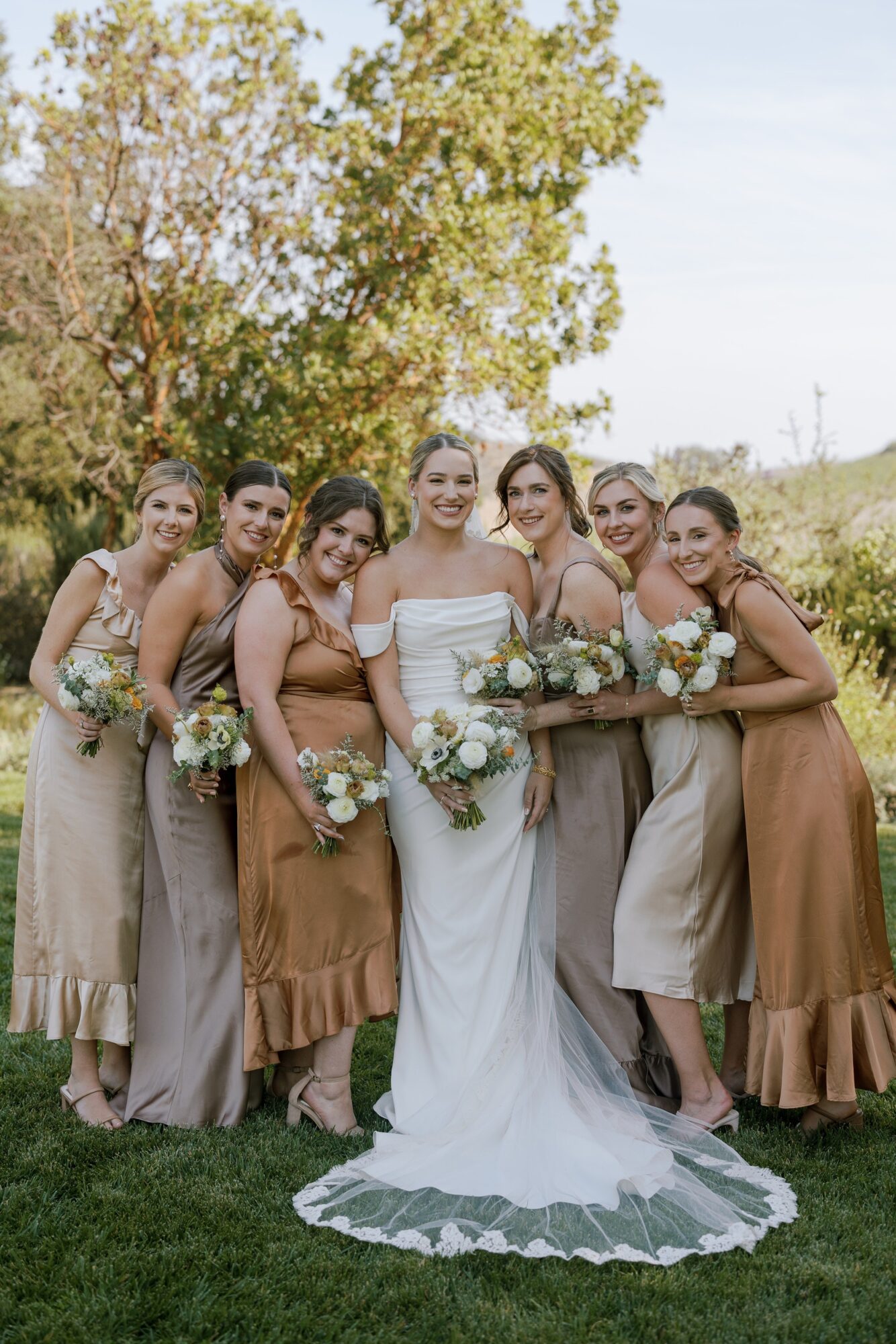 Group of seven women in wedding attire outdoors, holding bouquets, standing on grass with trees in background.