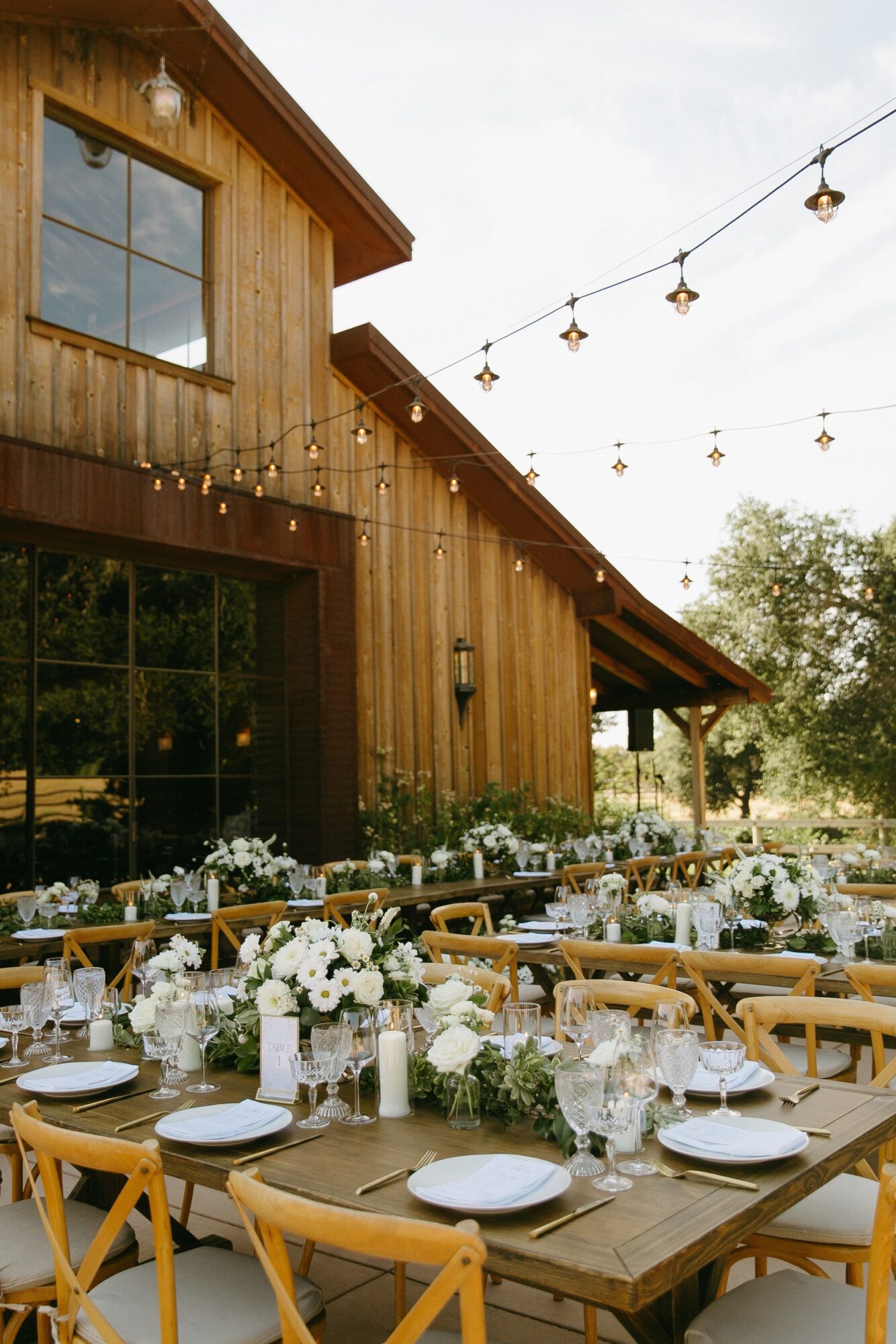 Outdoor dining area with tables, chairs, and floral centerpieces in front of a wooden building, string lights overhead, daytime setting.