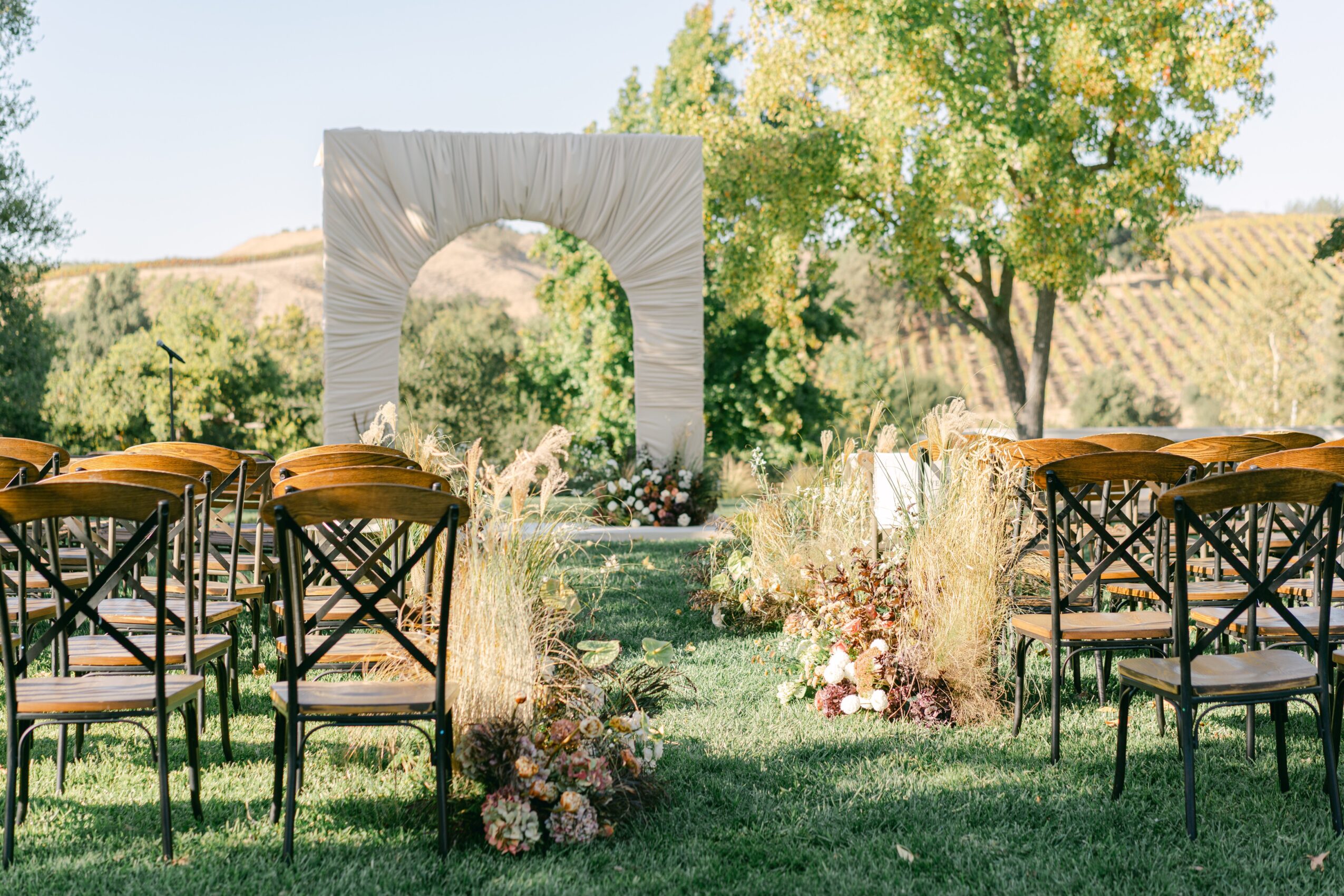Outdoor wedding setup with chairs facing a decorated arch and fountain, surrounded by greenery and trees.