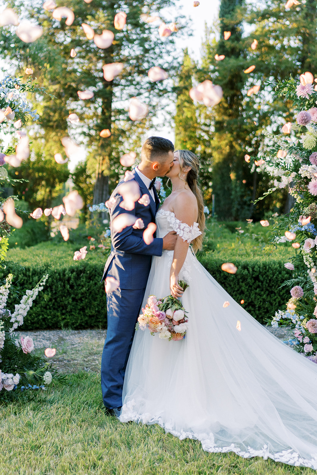 A bride and groom kiss outdoors surrounded by pink and white flowers, trees, and greenery, during daytime.