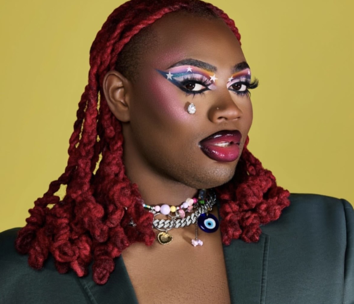 Woman with vibrant makeup, red braided hair, and colorful jewelry against a yellow background.