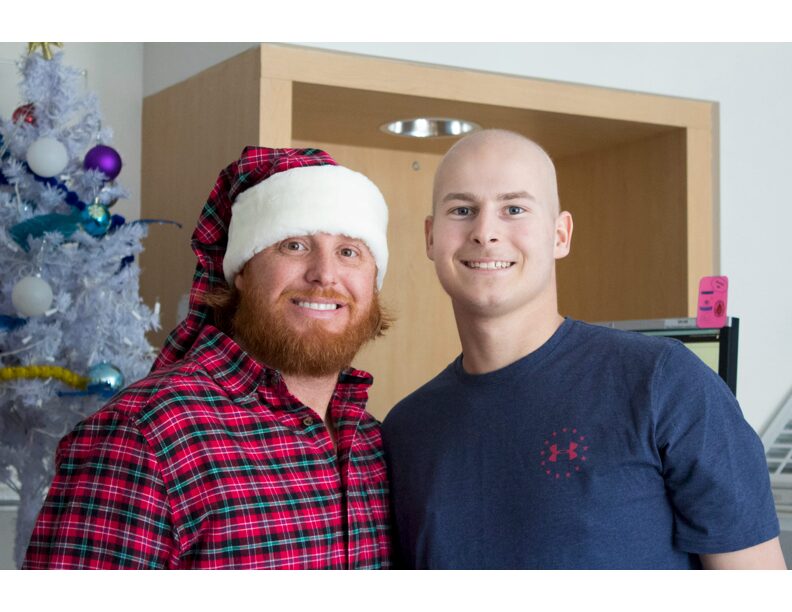 Two men smiling indoors, one wearing a Santa hat and plaid shirt, the other in a blue shirt, with a decorated Christmas tree nearby.