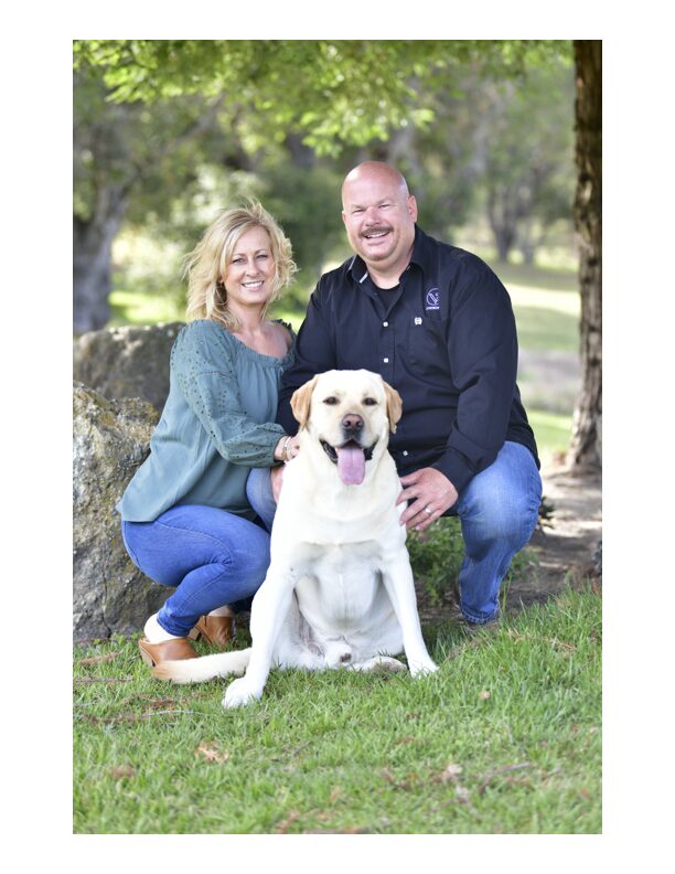 A smiling woman, man, and a Labrador Retriever dog outdoors in a park with trees and grass.
