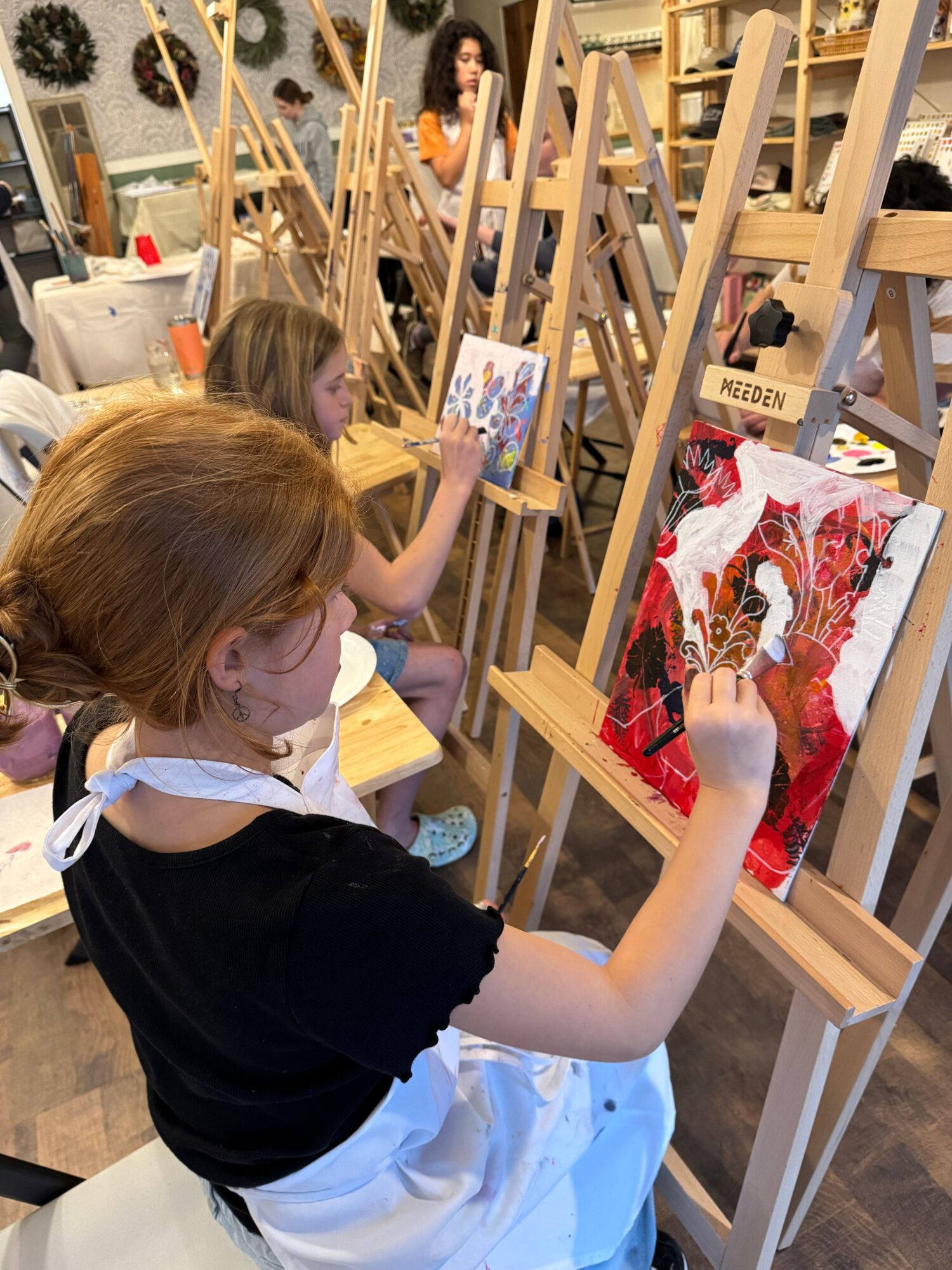 Child with red hair painting on a canvas at an art display, surrounded by wooden easels and art supplies.