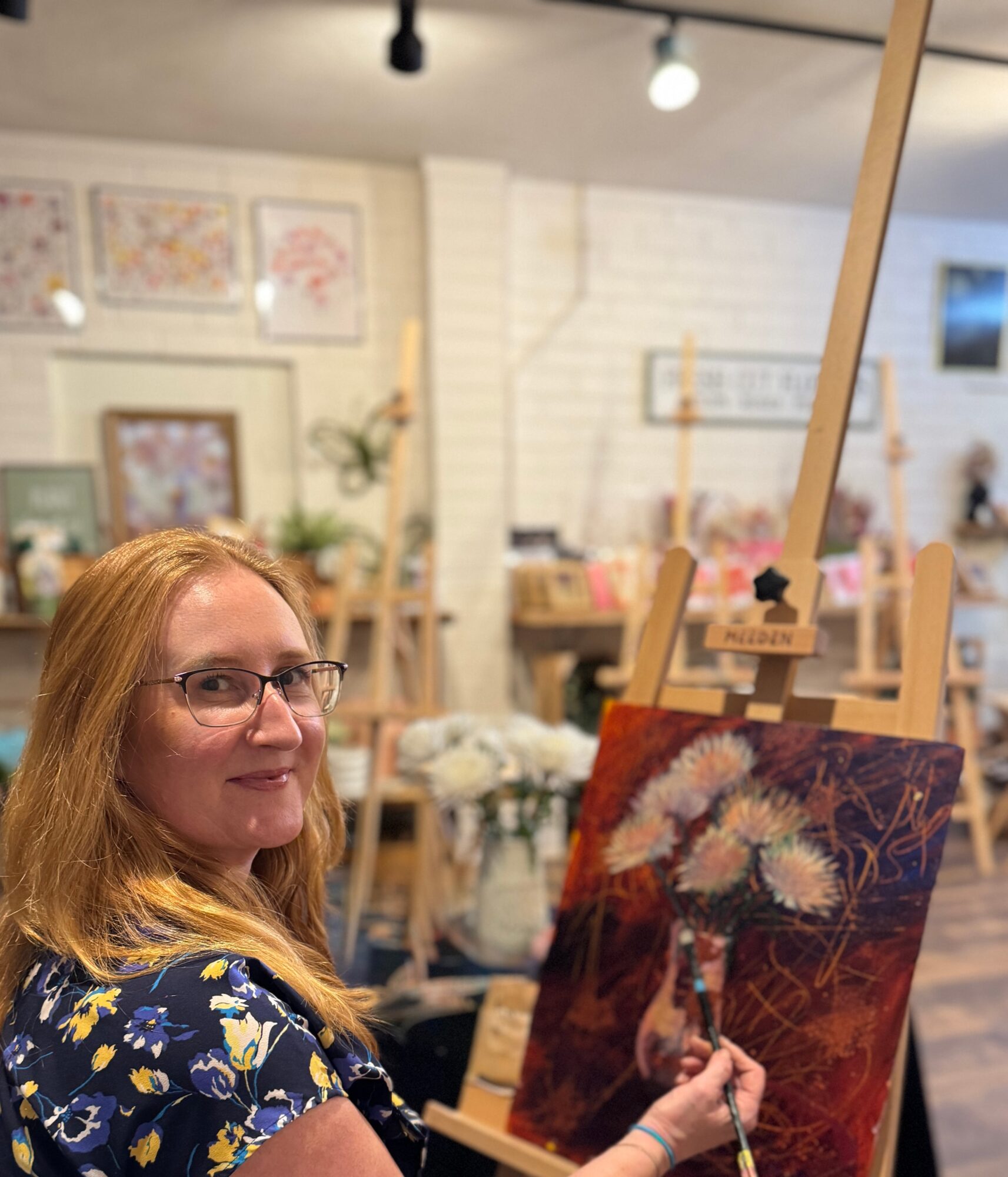 Woman with glasses and long hair painting a floral still life on an easel in a cozy art studio.