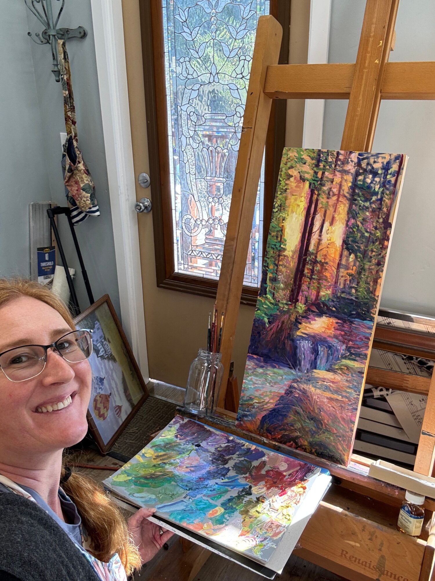 Person with glasses smiling, surrounded by colorful paintings and art supplies in a room.