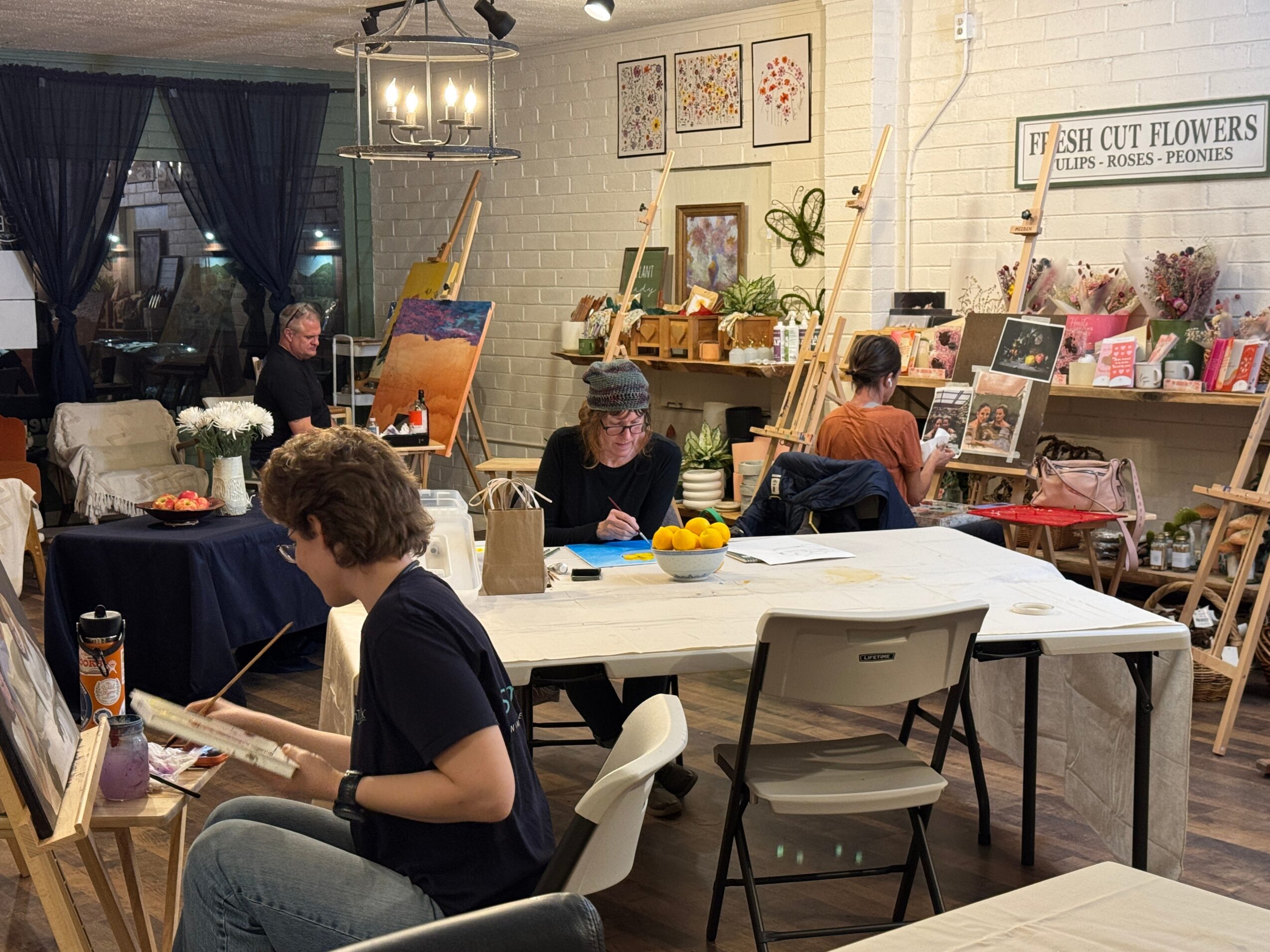 People painting and browsing art supplies in a cozy art studio with white brick walls and shelves of flowers and art materials.
