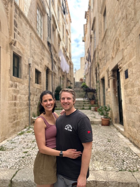 A smiling couple stands close together in a narrow stone alleyway with buildings on both sides, stairs, and potted plants.