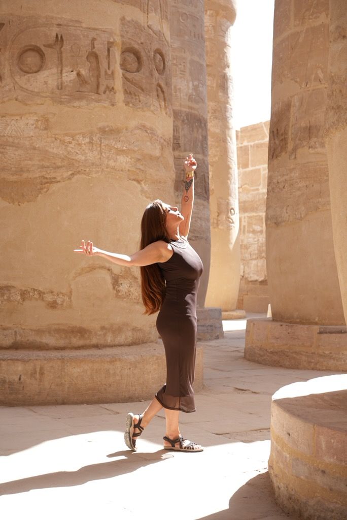Woman with long hair in a dress and heels raises arms in an ancient stone structure, sunlight streaming in.
