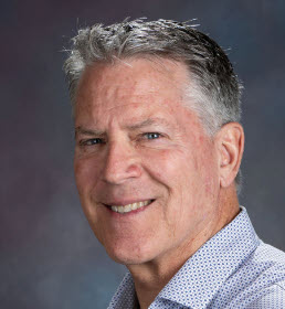 Portrait of a smiling middle-aged man with gray hair, wearing a light blue patterned shirt, against a dark background.