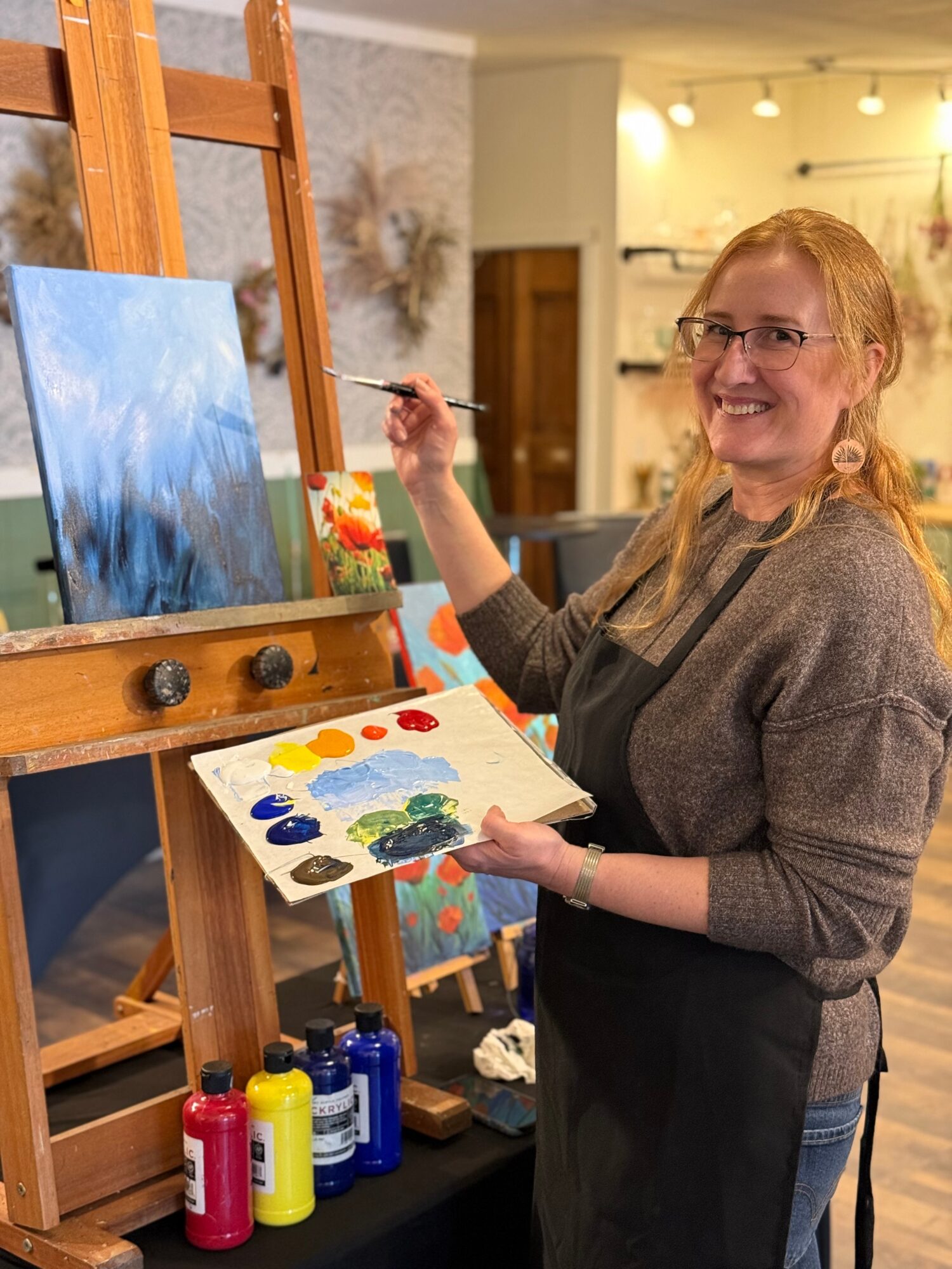 Woman with glasses smiling, holding a paintbrush and palette, standing next to an easel with a painting, in a cozy indoor space.