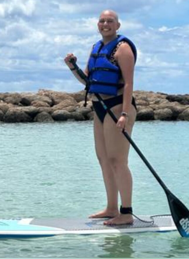 Person standing on paddleboard in water, wearing a life jacket, holding a paddle, with rocks and sky in background.
