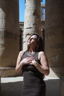 Woman standing outdoors near ancient stone columns, wearing a black dress, with her hand on her chest, smiling.