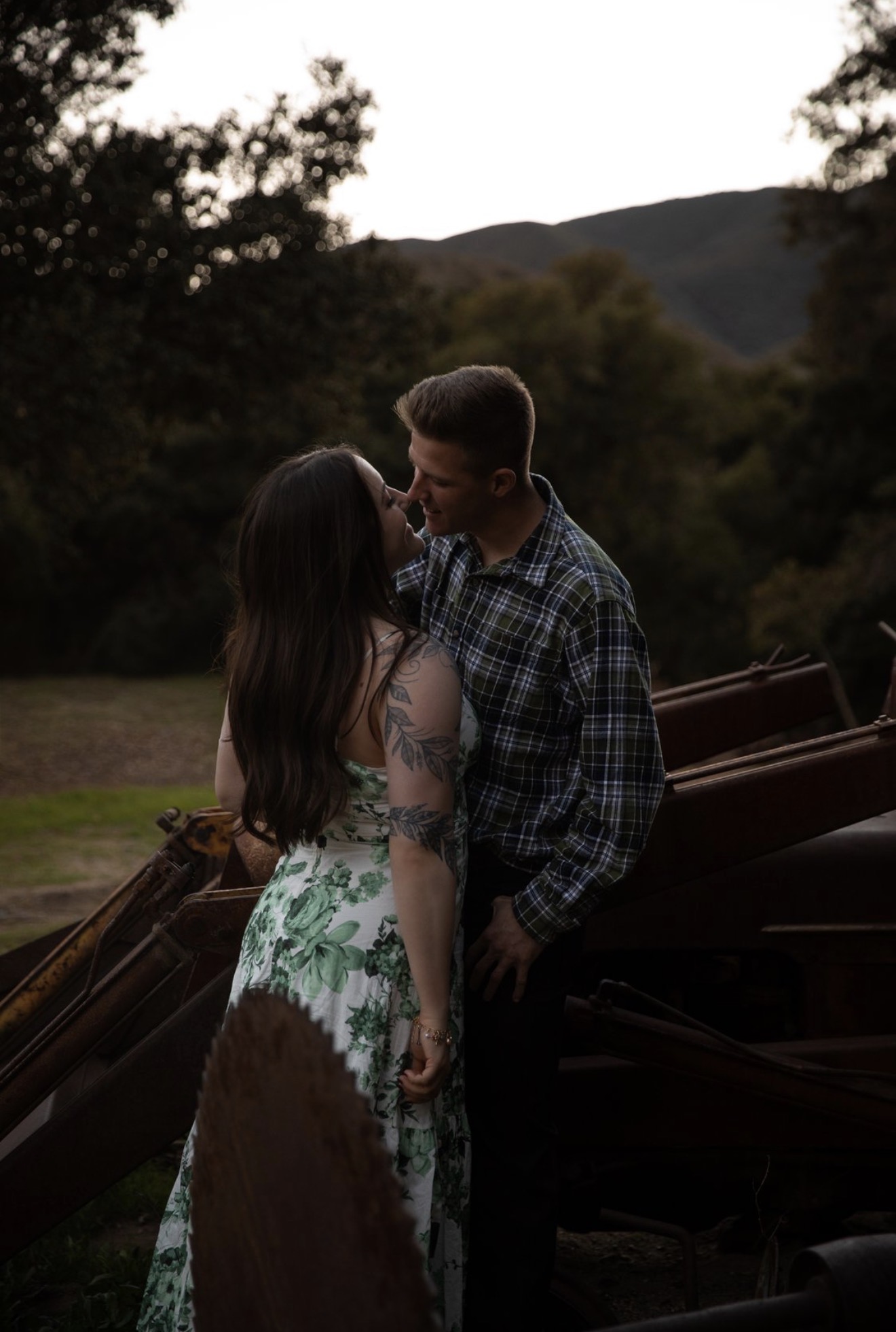 A couple stands close outdoors with trees and hills in the background, sharing an intimate moment.