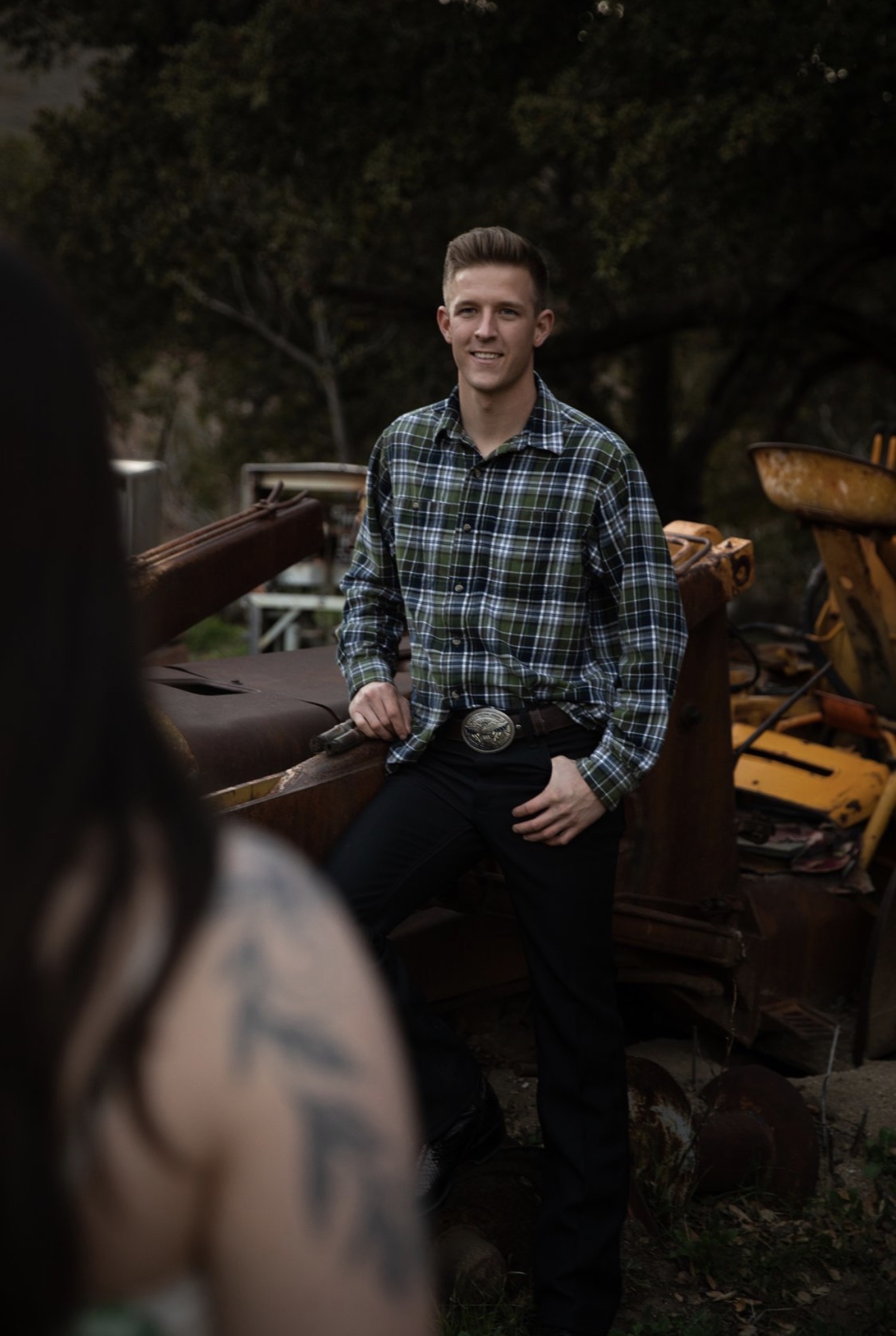 Young man in plaid shirt standing outdoors with trees and equipment in background.