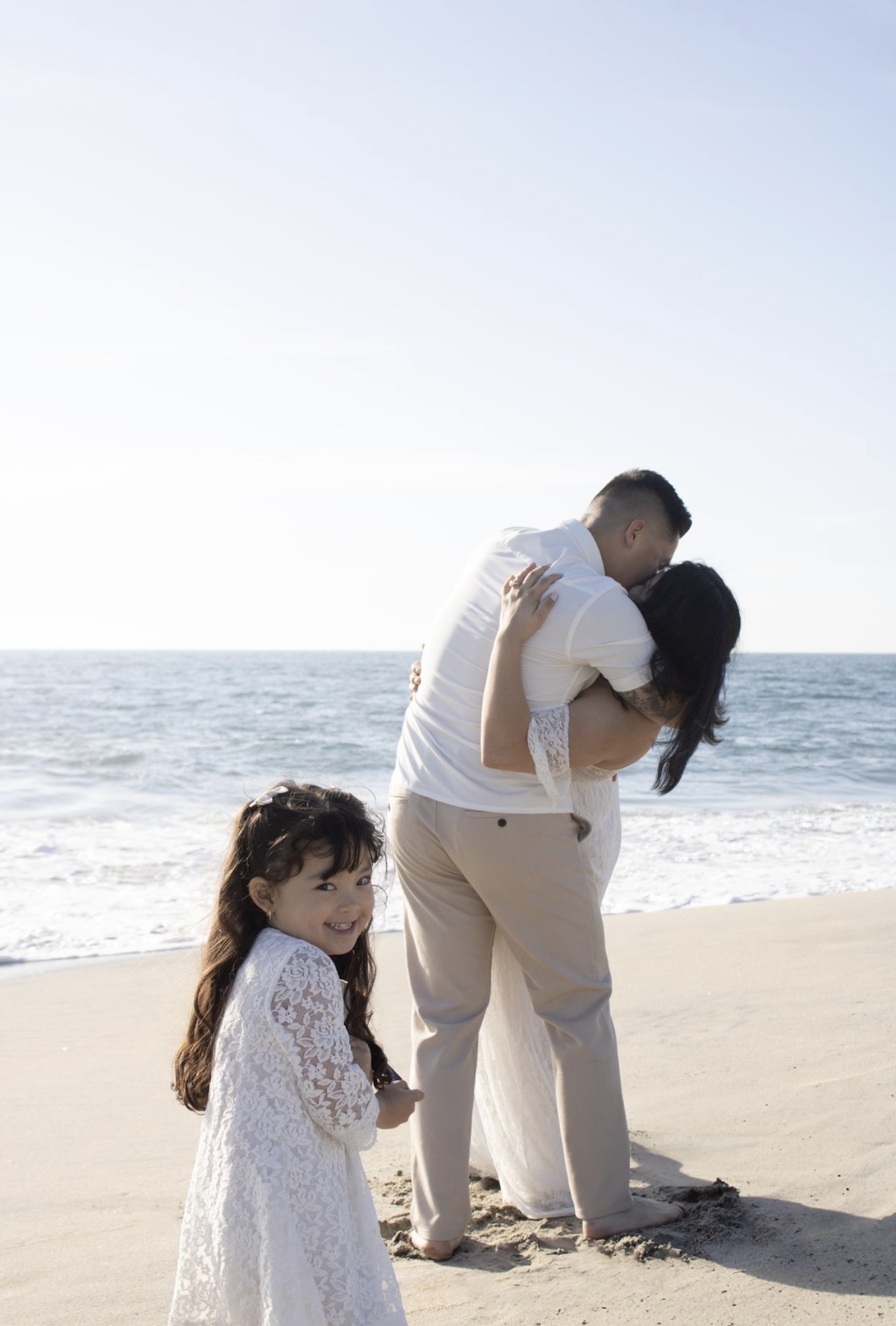 A couple embraces on the beach with a young girl smiling nearby, ocean and sky in the background.