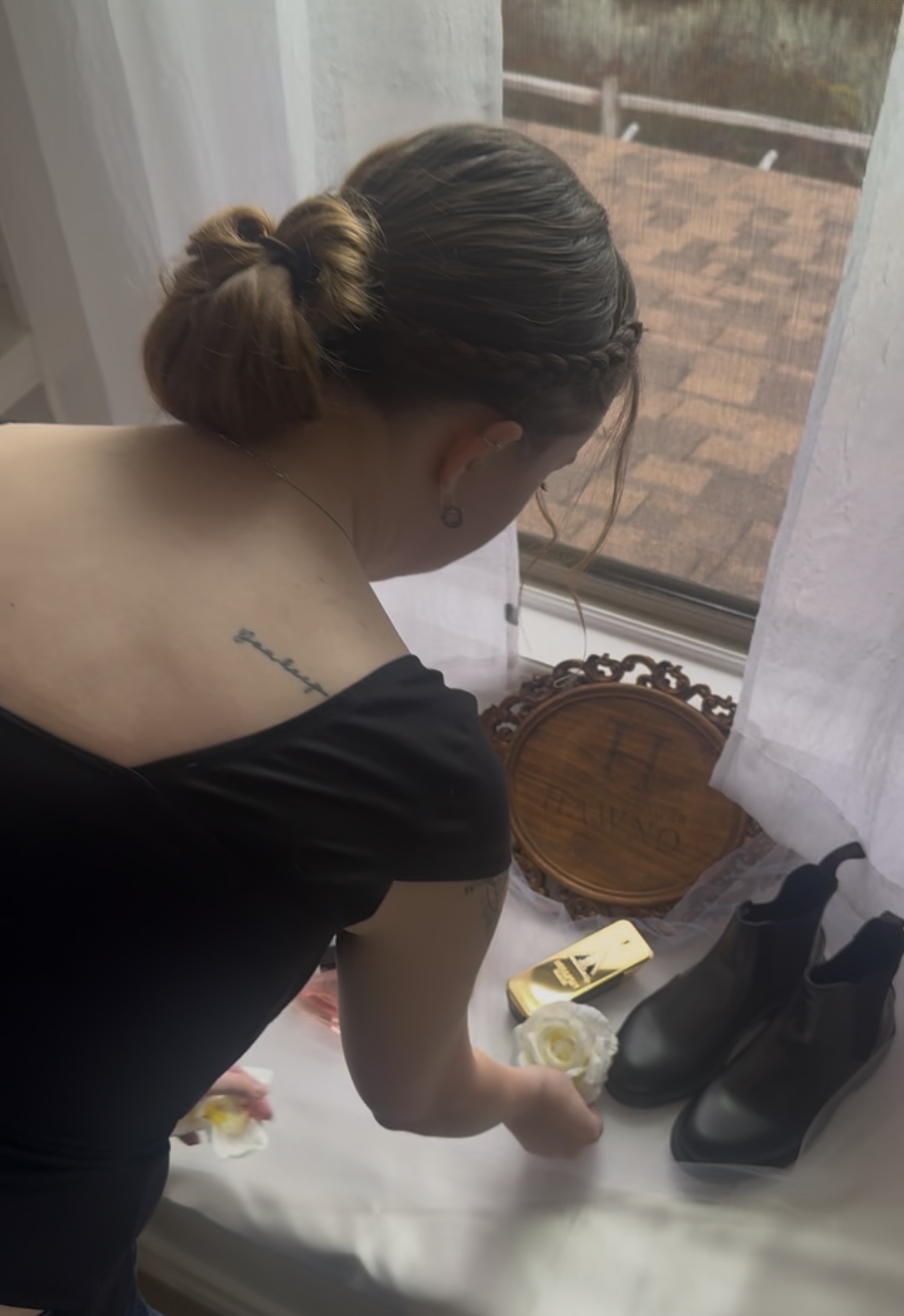 Person with hair in bun reaching for shoes on a windowsill, with a tattoo on shoulder visible, outside view through window.