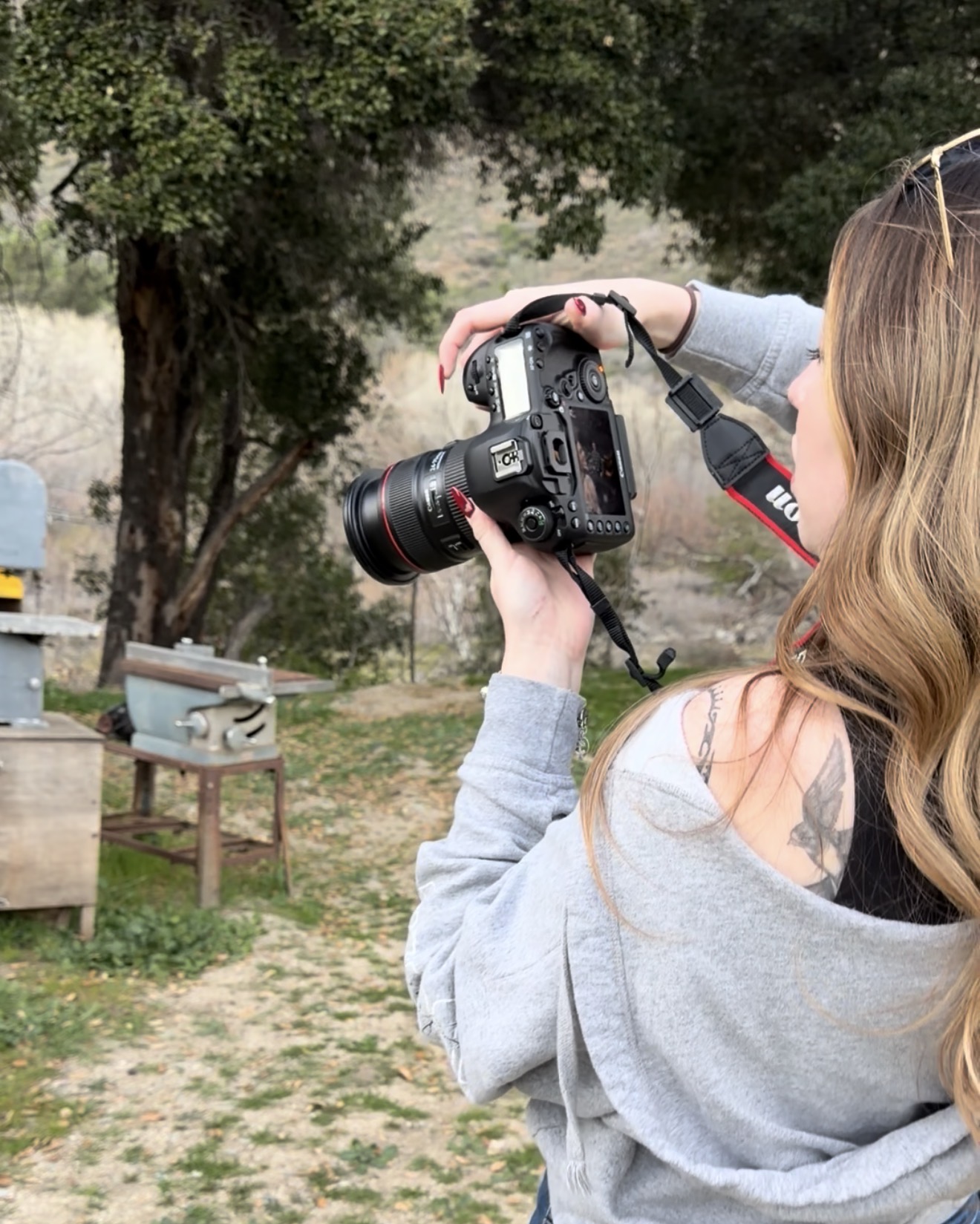 Person with long hair holding a camera outdoors, trees and a bench in the background.