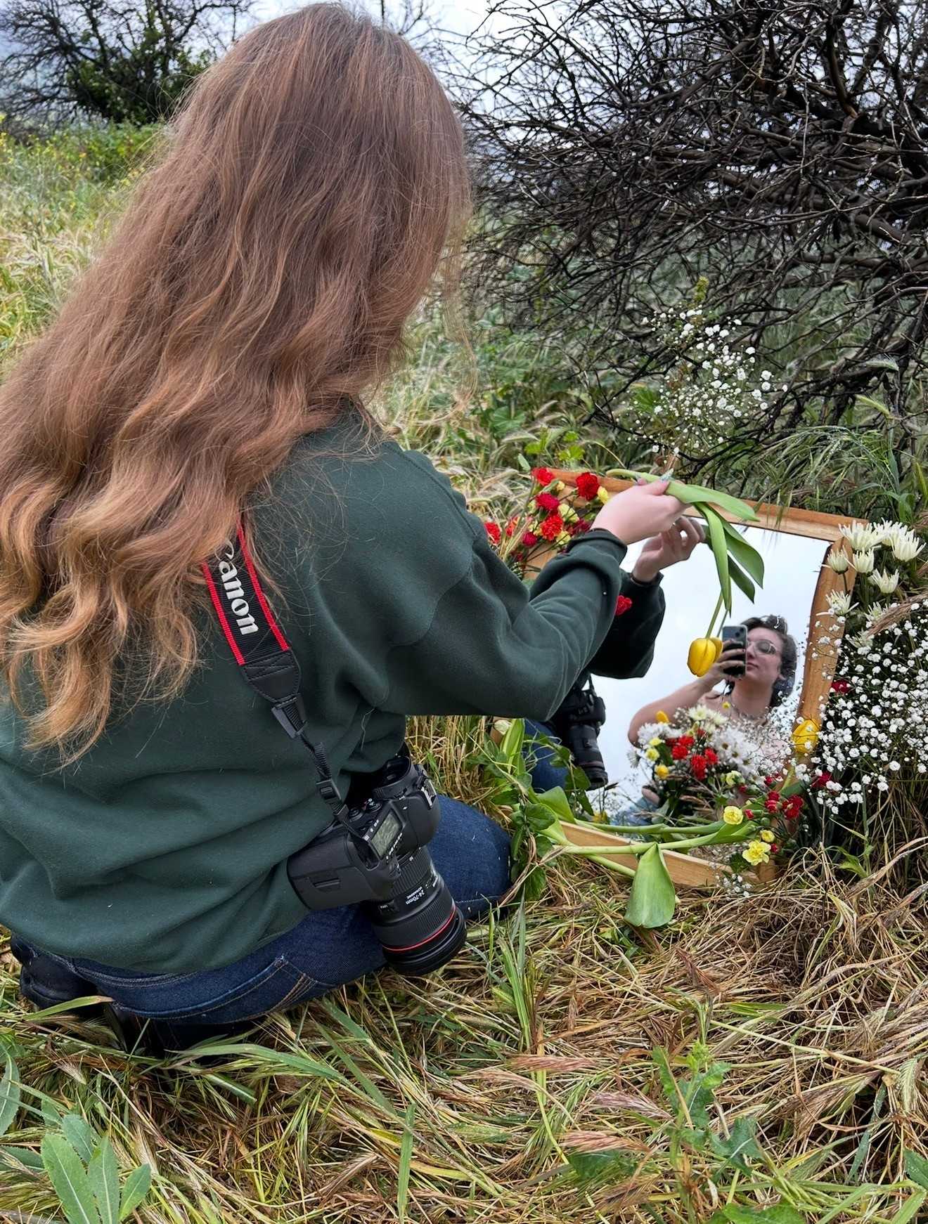 Person with long hair placing flowers at a memorial with a mirror reflecting their face, outdoors with grass and trees.