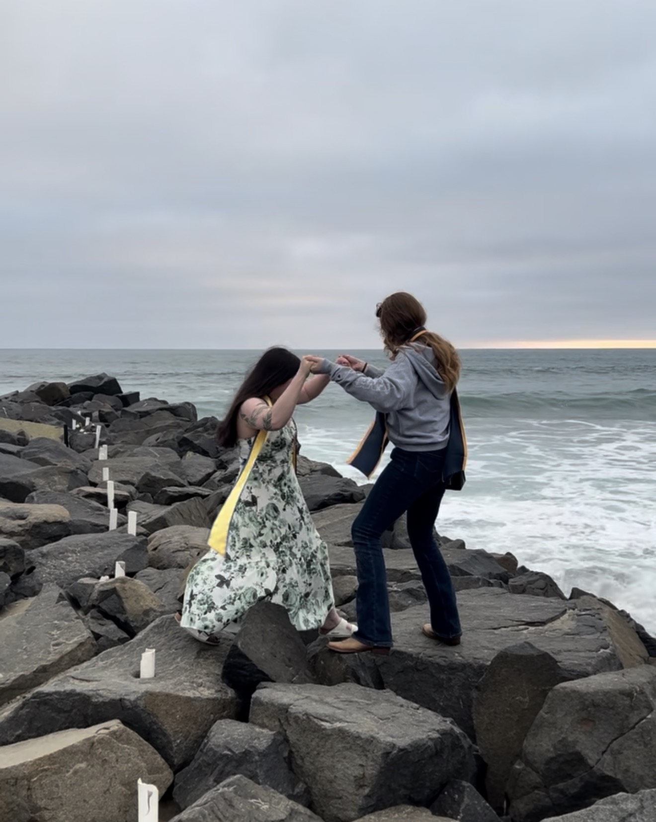 Two women dancing on rocks by the ocean under cloudy sky, holding hands and facing each other.