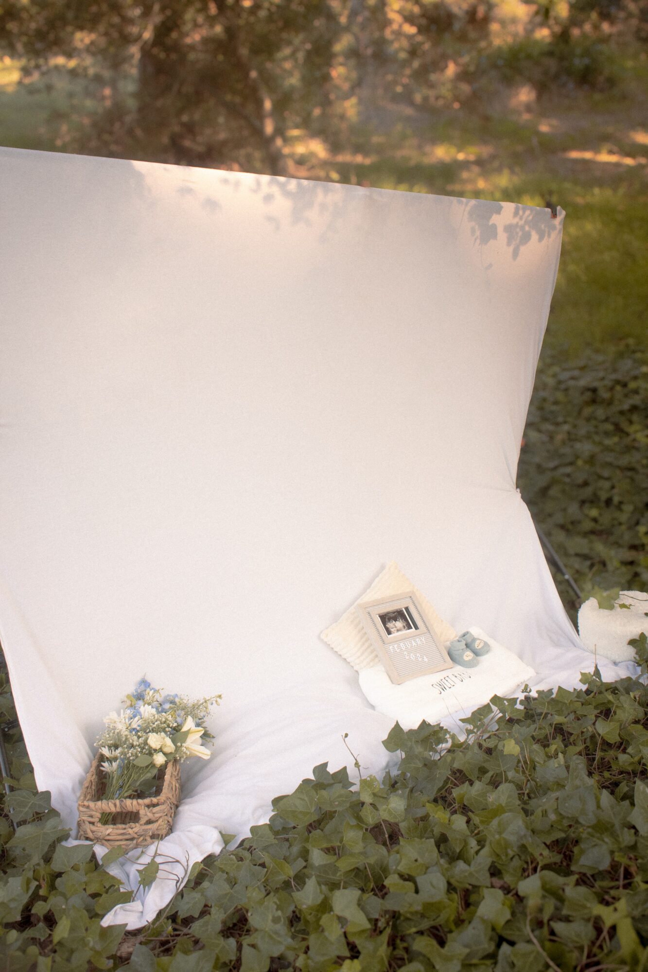 Outdoor scene with a white backdrop, a basket of flowers, and a vintage telephone on a white cloth, surrounded by greenery.