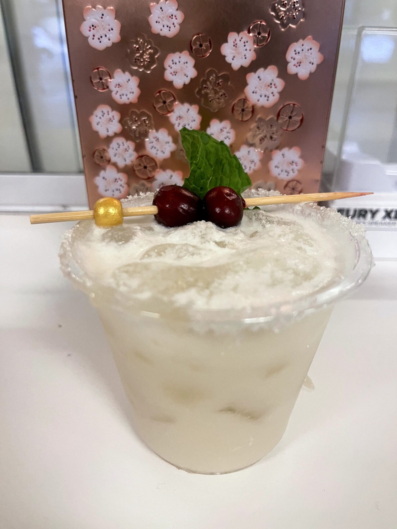 Creamy dessert in a glass with cherries, mint, and a decorative stick, with a patterned background behind.