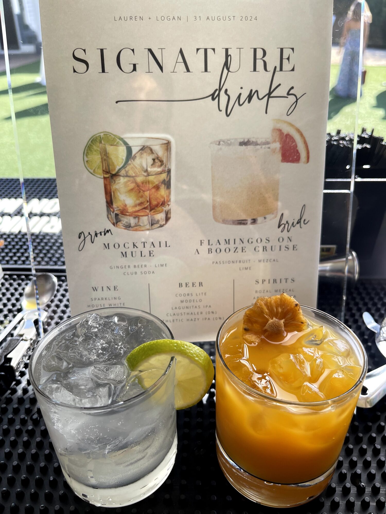 Two drinks on a table with a menu behind them, one clear with lime and ice, the other orange with a flower garnish.