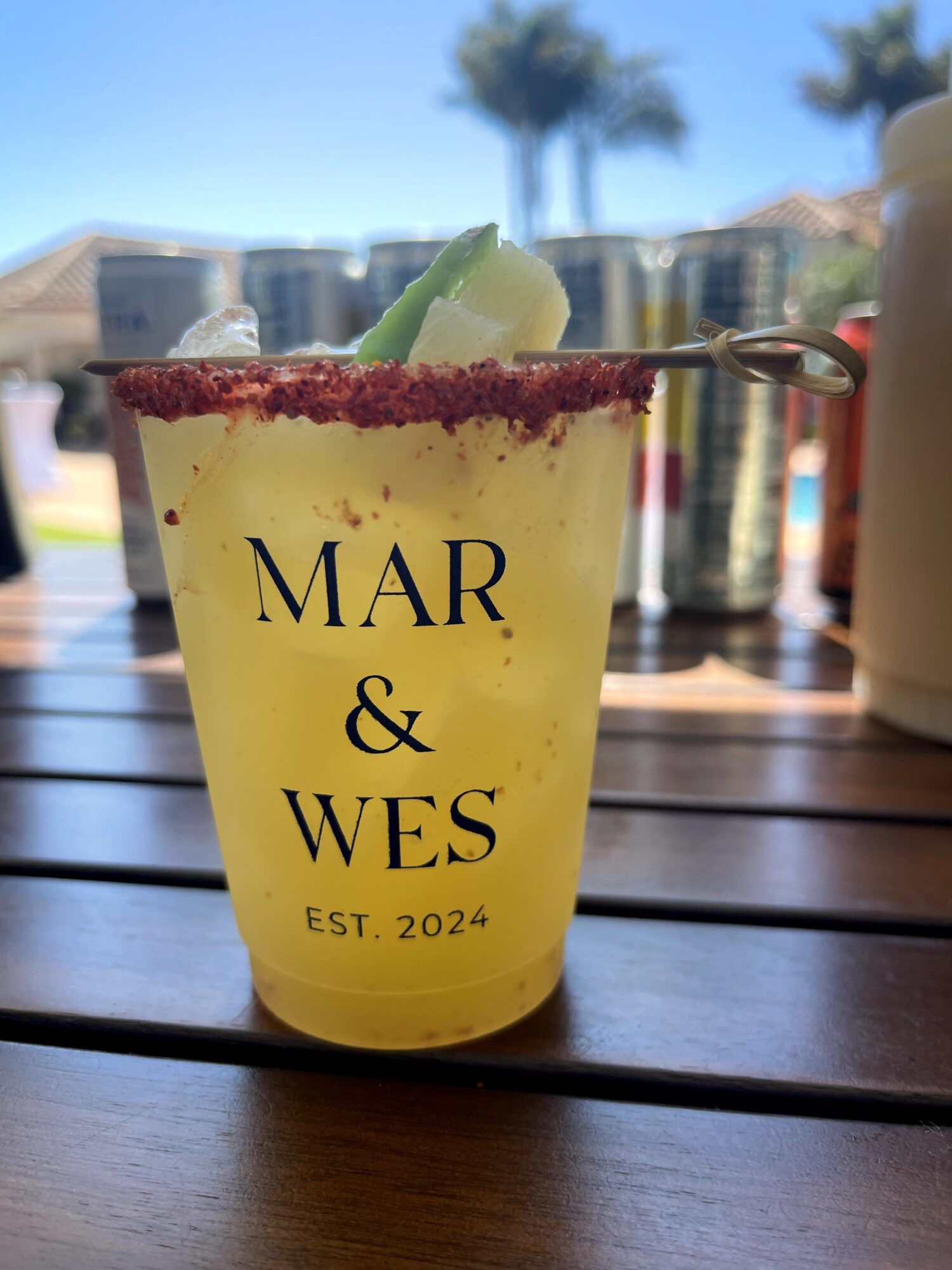 Yellow cocktail in a clear cup with lime wedge, set on a wooden table outdoors with palm trees in background.