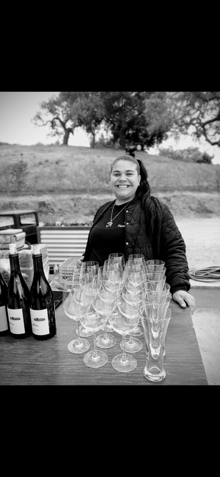 Smiling woman standing behind a table with wine bottles and glasses outdoors, trees in background.