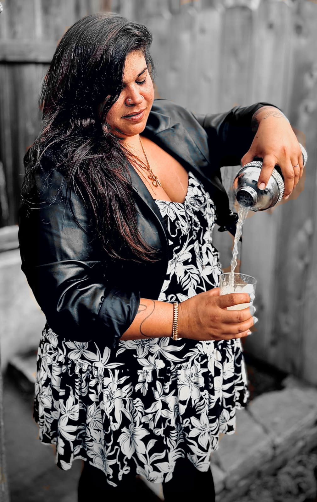 Woman pouring a drink into a glass outdoors, wearing a black leather jacket and patterned dress, with a wooden fence background.