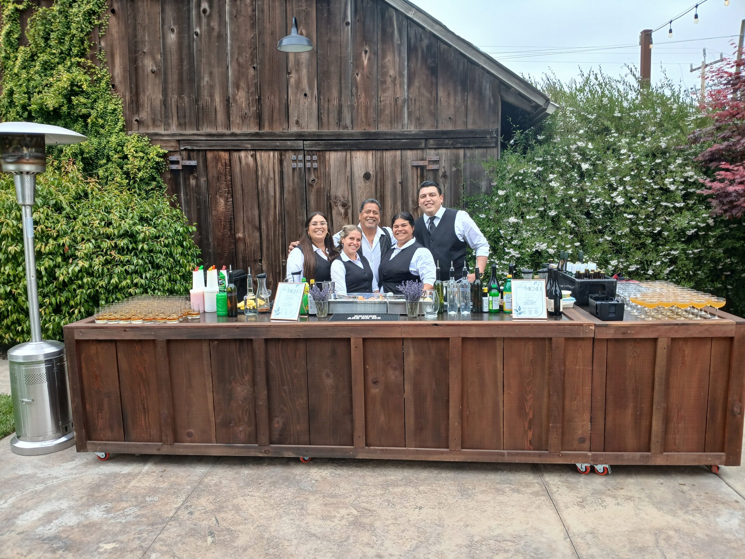 Group of five people behind a wooden bar outdoors, with greenery and a barn in the background.