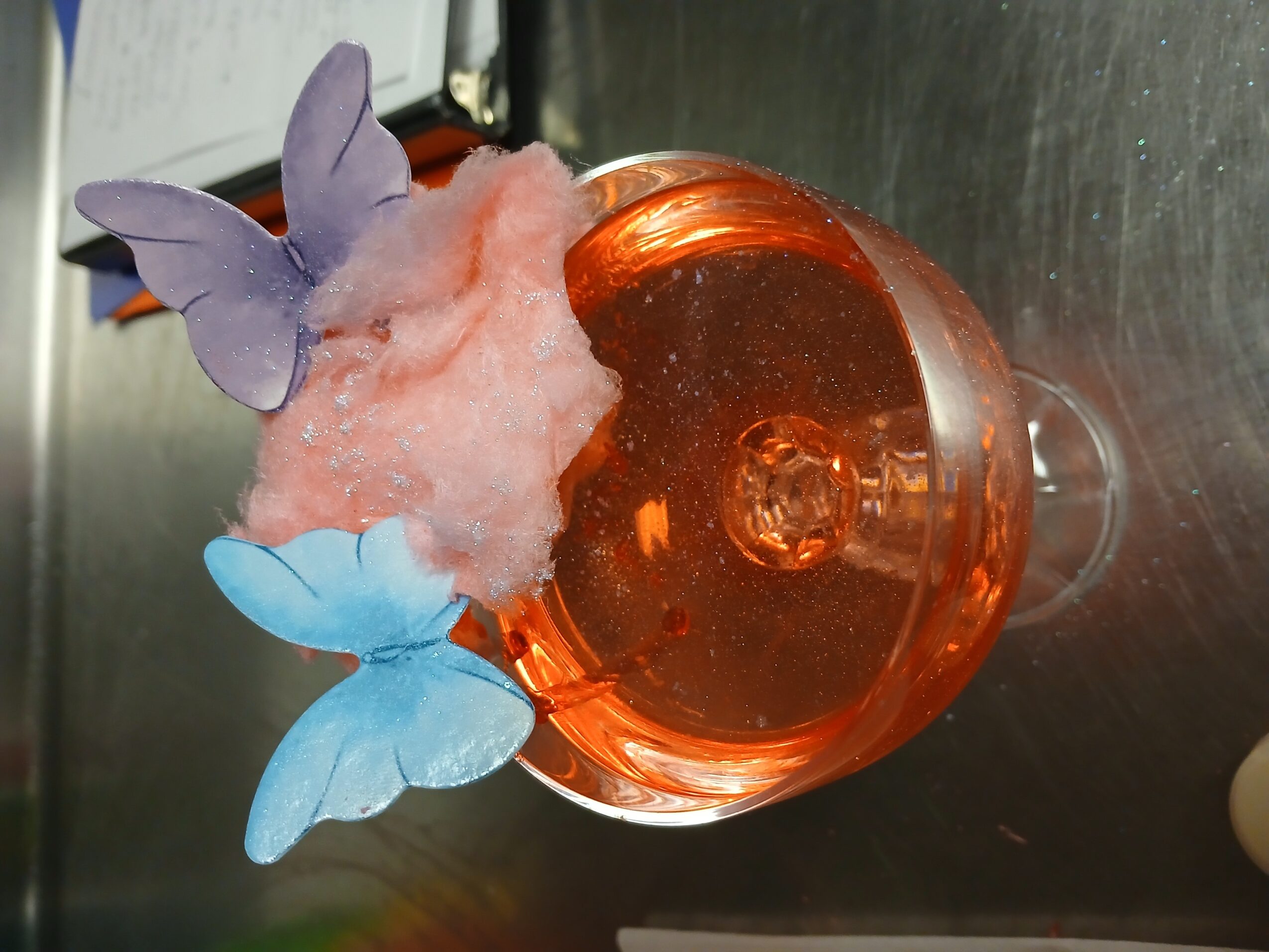 Glass with pink cotton candy and blue butterfly decorations, viewed from above on a metallic surface.