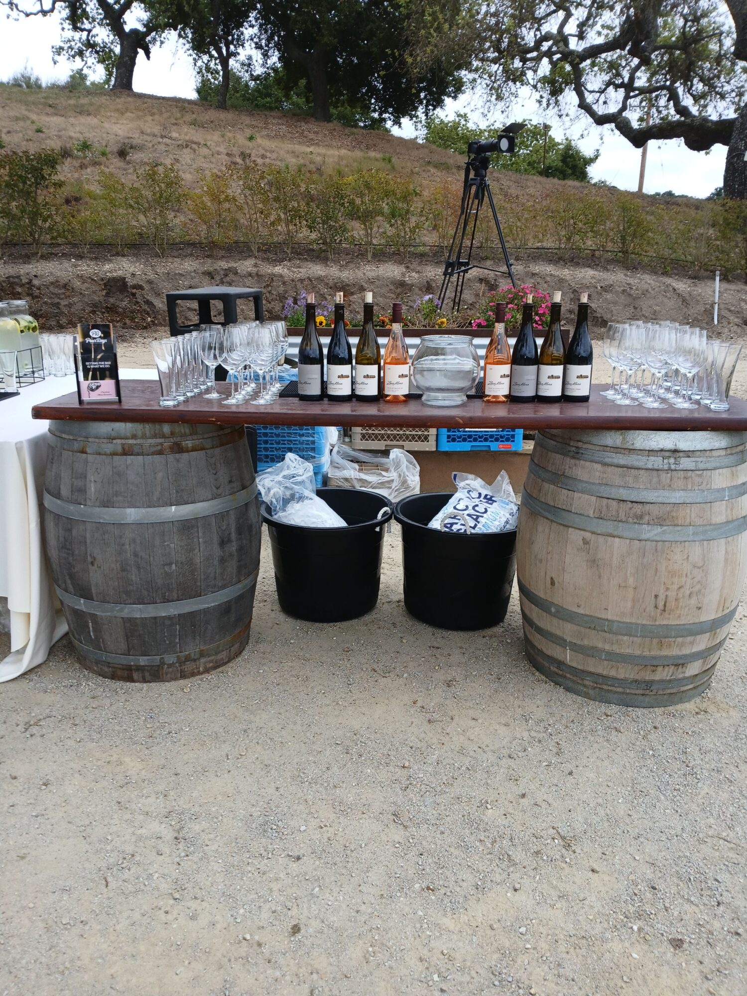 Outdoor setup with barrels, bottles, and supplies on a table, outdoors with trees in the background.