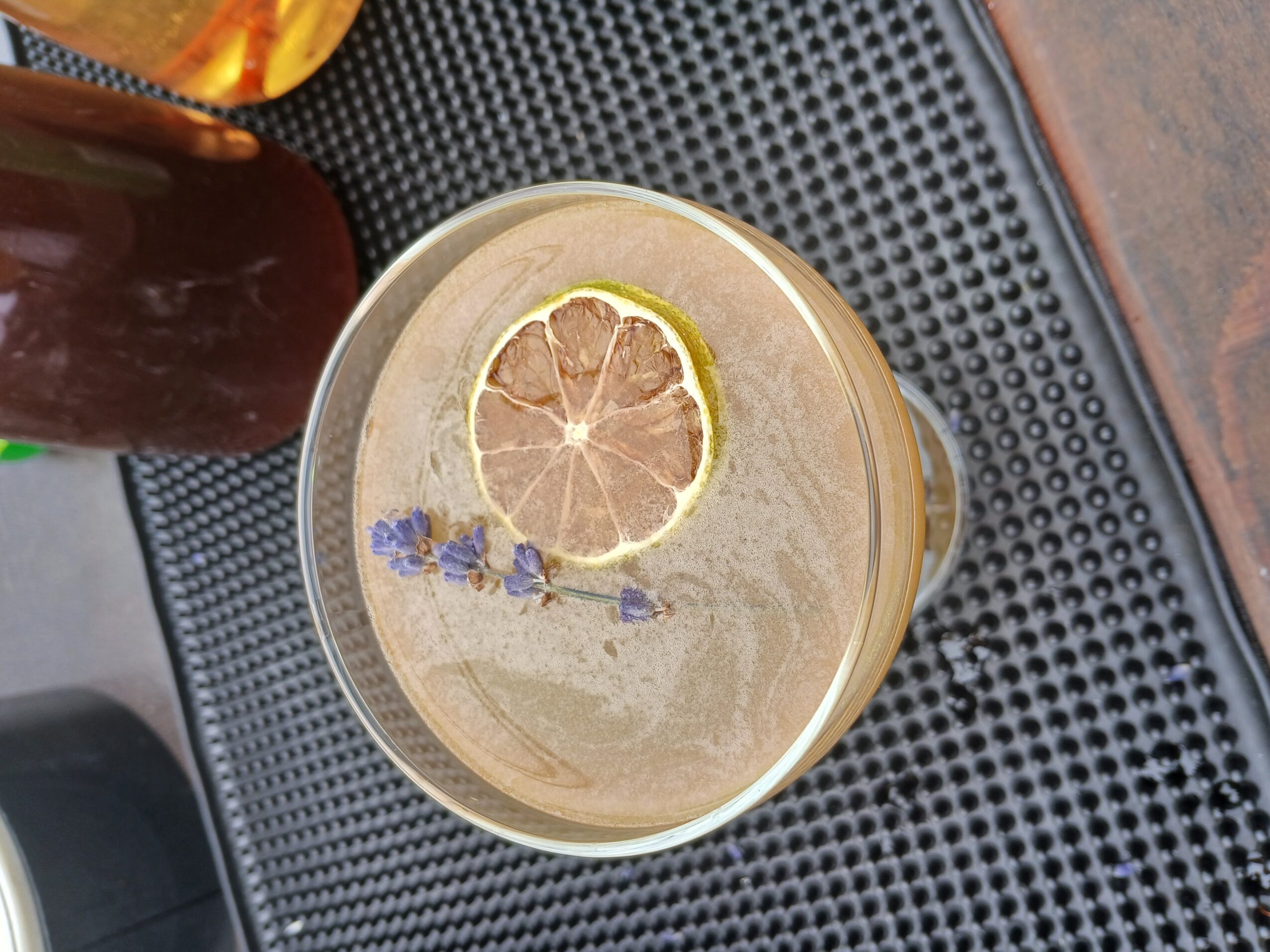 A glass of beverage with lemon slice and lavender on top, placed on a black textured surface.