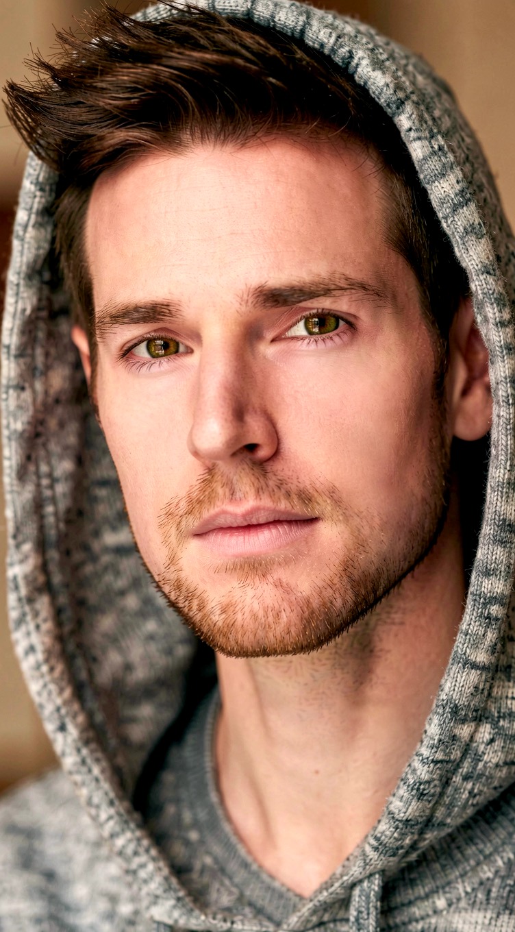 Close-up of a young man with short dark hair, wearing a gray hoodie, looking directly at the camera.