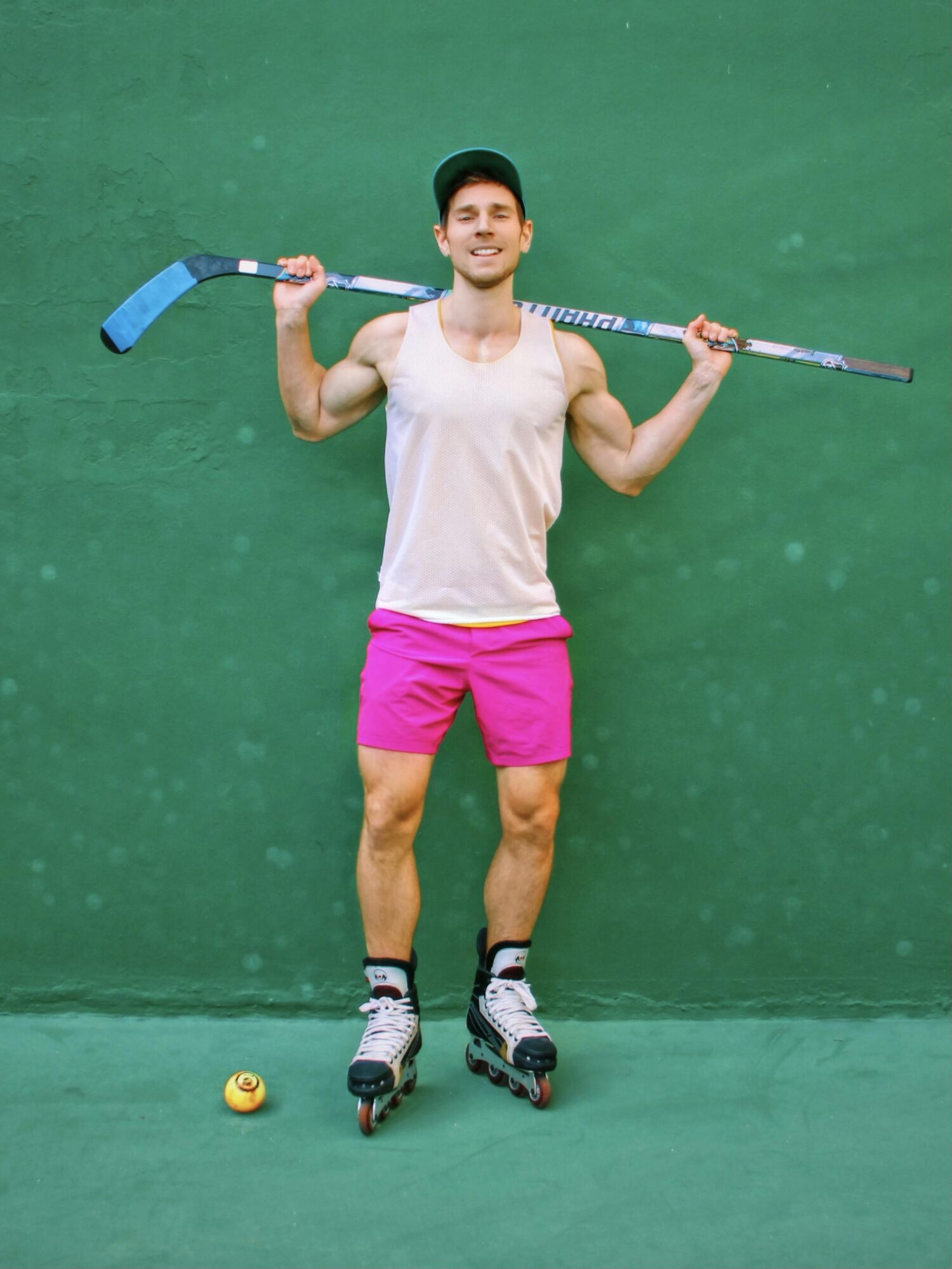 Young man in pink shorts and a white tank top on roller skates holding a hockey stick, standing on green background.