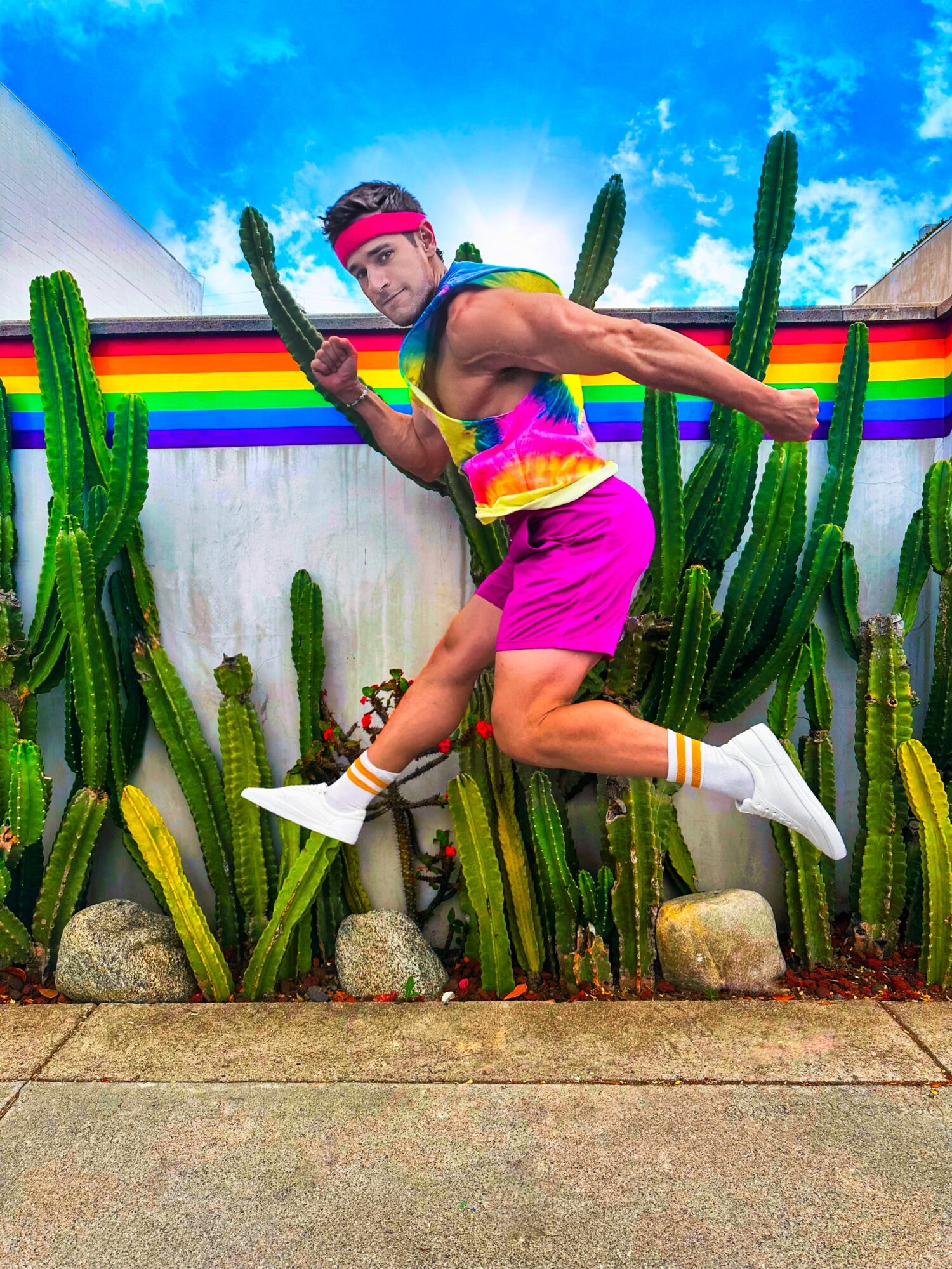 Person jumping among tall cacti with a rainbow-colored wall and blue sky background, wearing colorful clothing and white shoes.