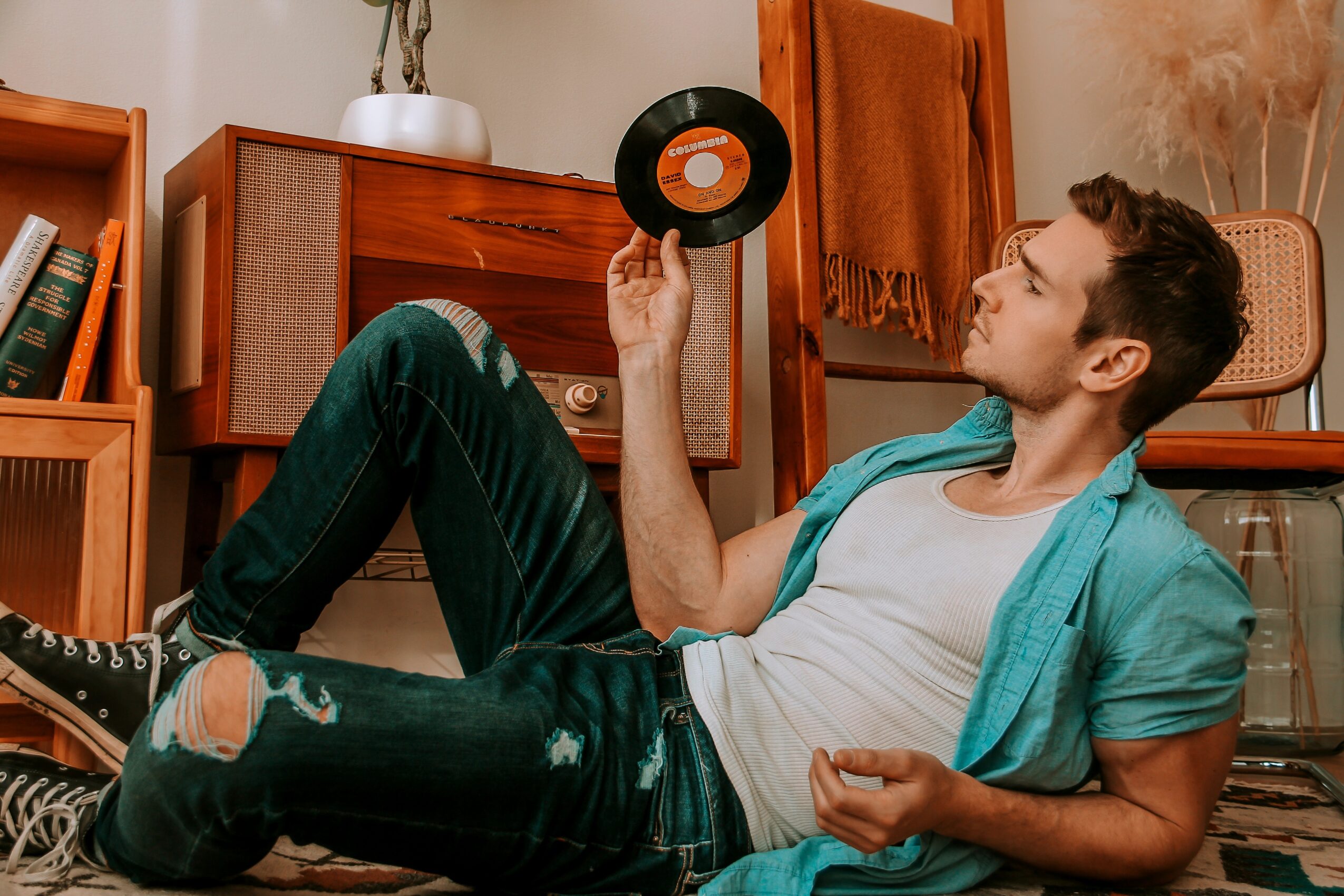 Young man lying on the floor, holding a vinyl record, with furniture and books in the background.