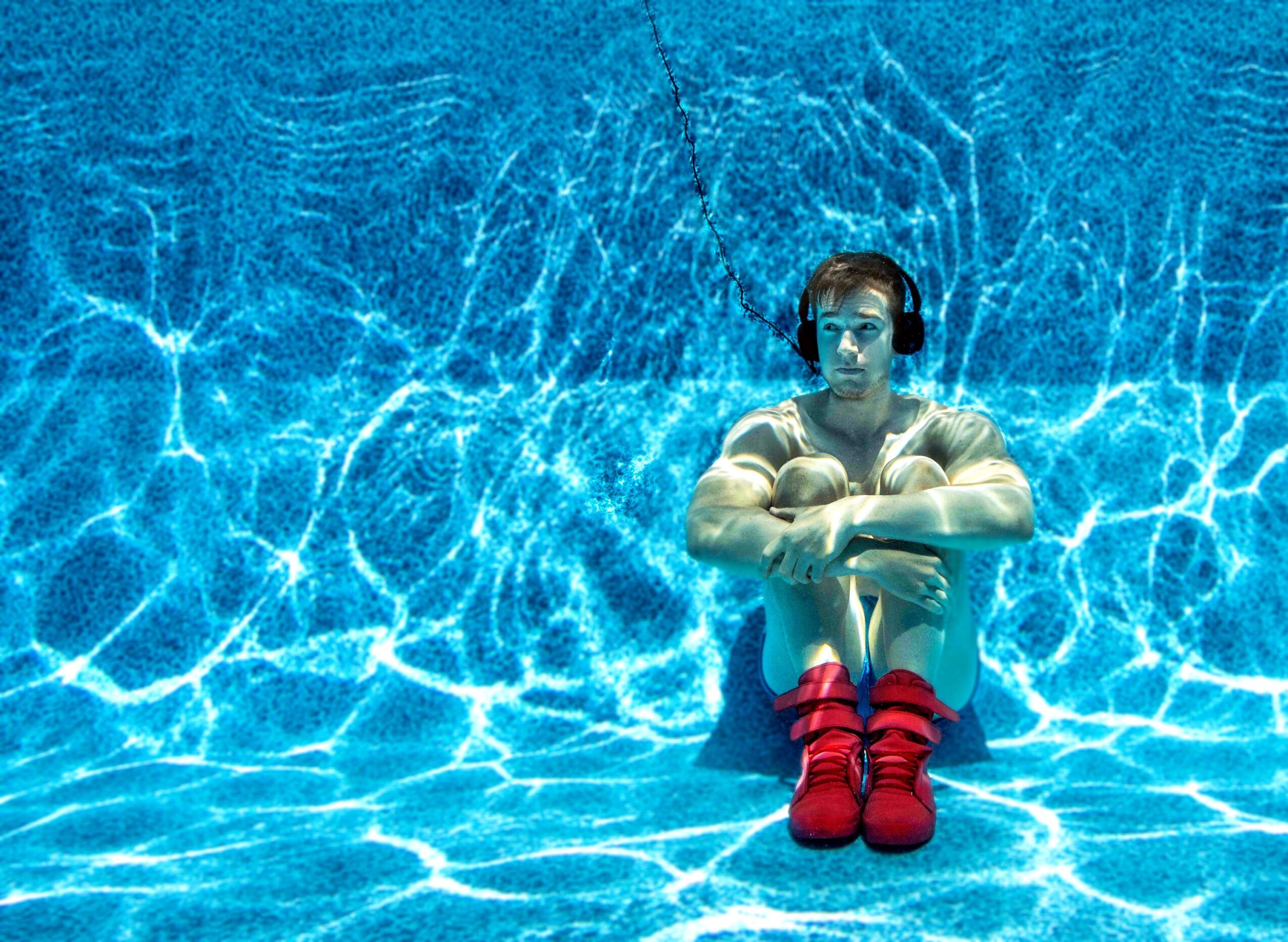 Person sitting underwater with arms crossed, wearing headphones and red shoes, surrounded by blue water reflections.