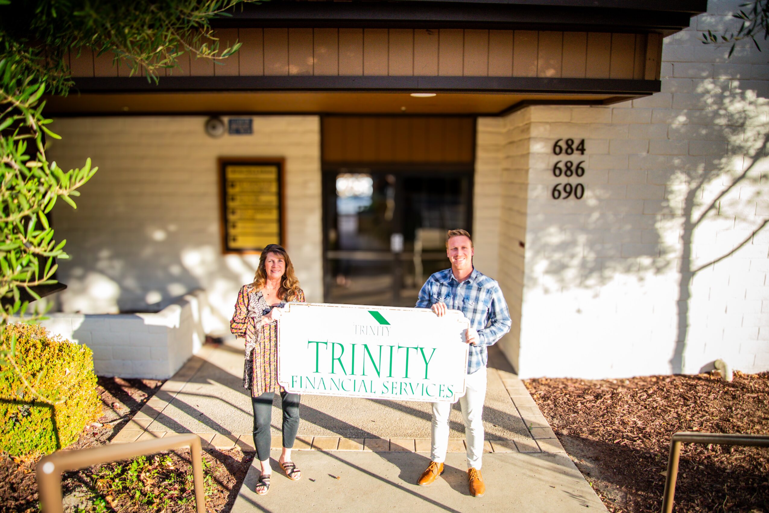 Two people holding a sign in front of a building entrance, with trees and bushes nearby.