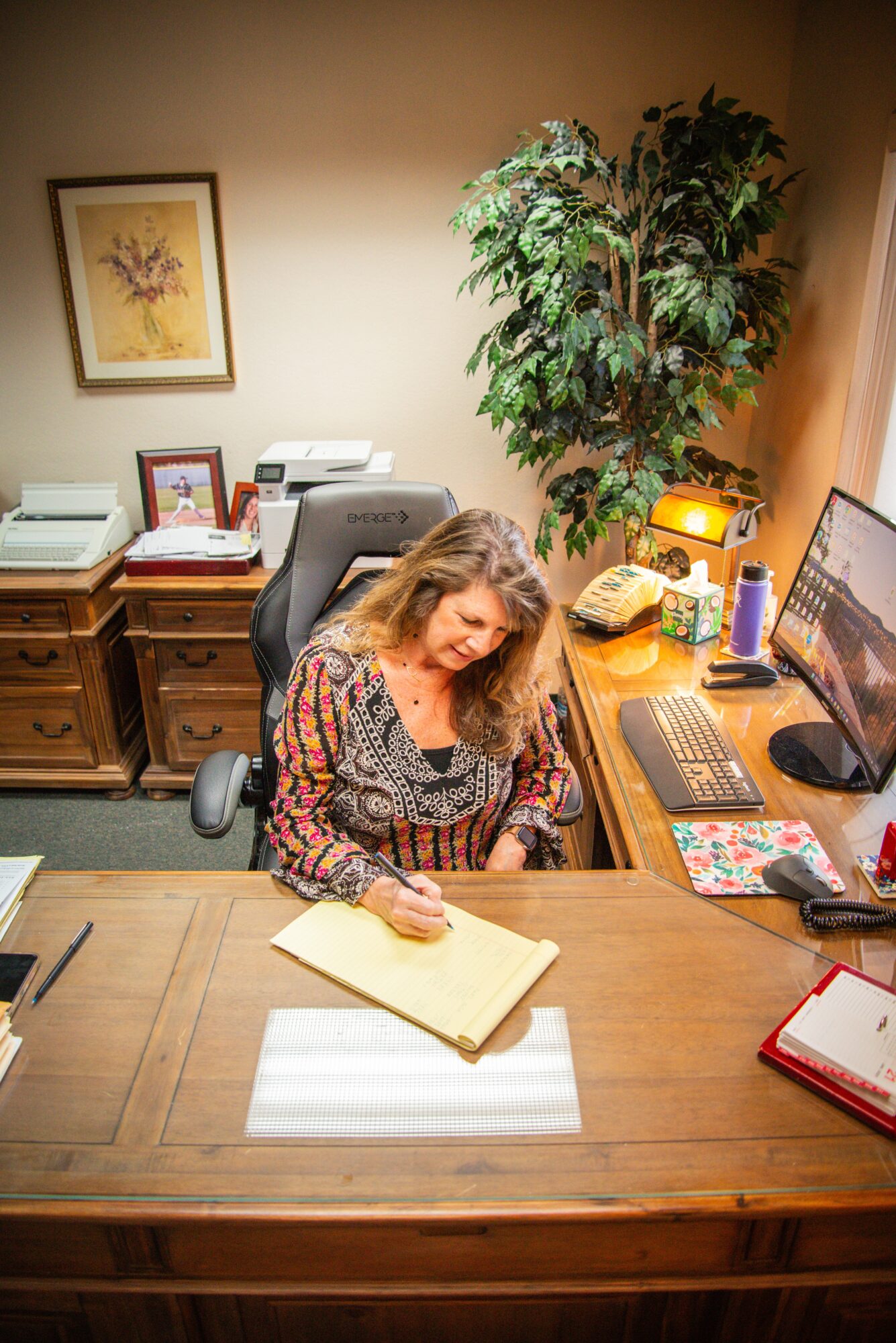 Woman sitting at a desk writing in a notebook, with a computer, lamp, and office supplies on the desk, and a large plant behind her.