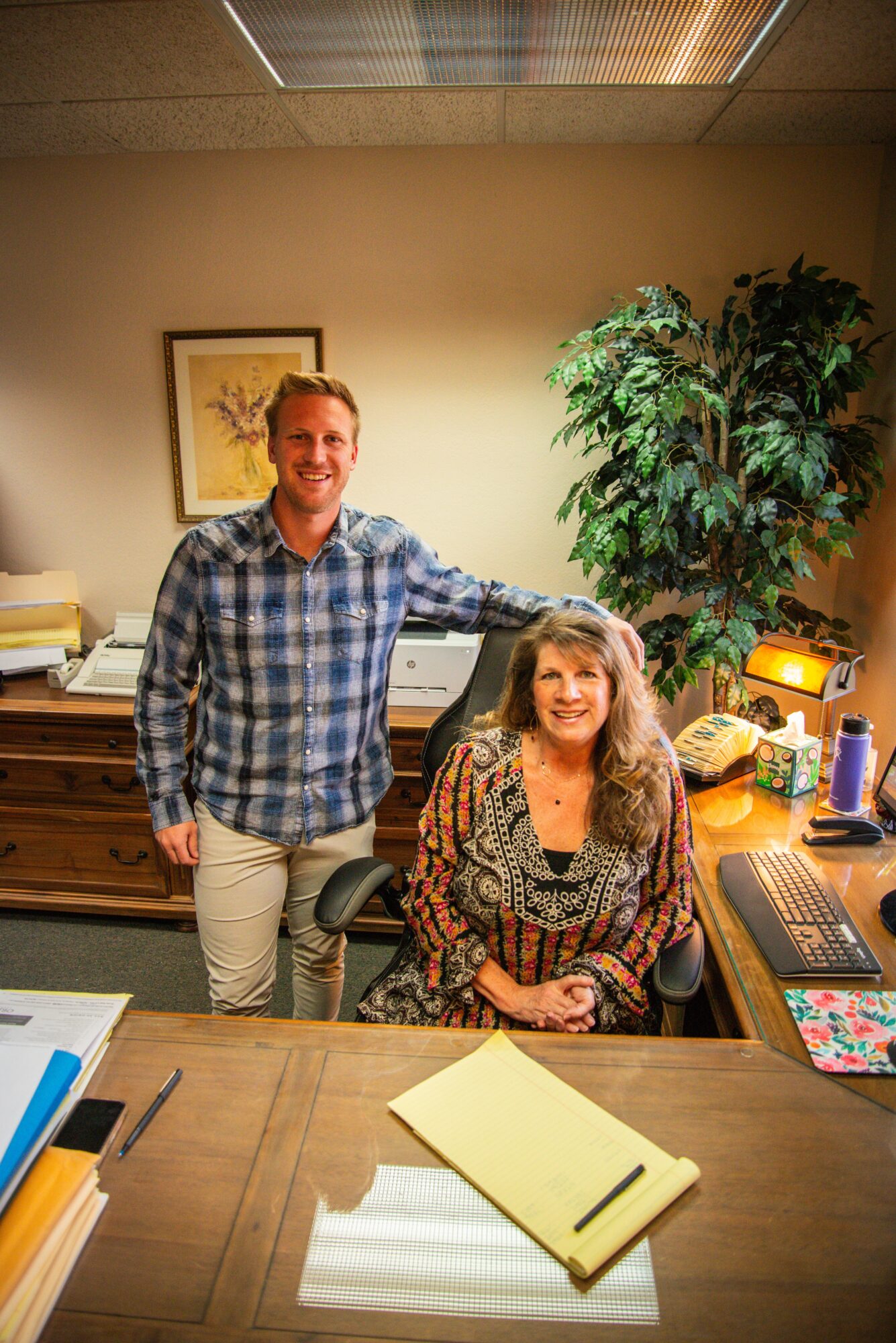 A man and a woman are in an office, smiling, with a large plant and framed picture behind them.