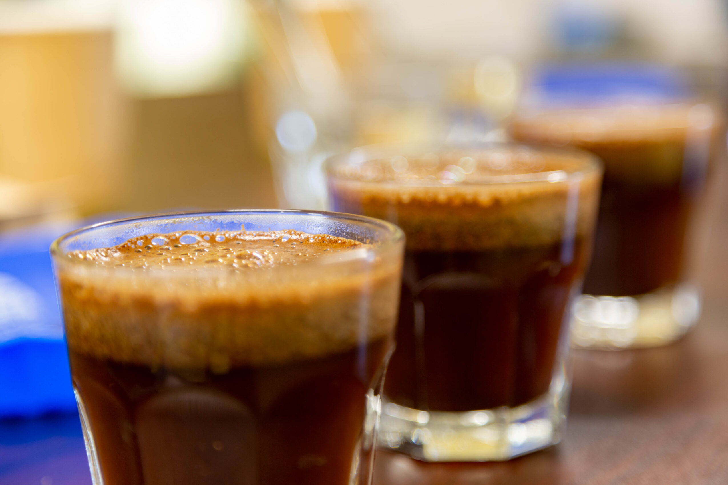 Three glasses of dark coffee with frothy tops on a wooden surface, blurred background.