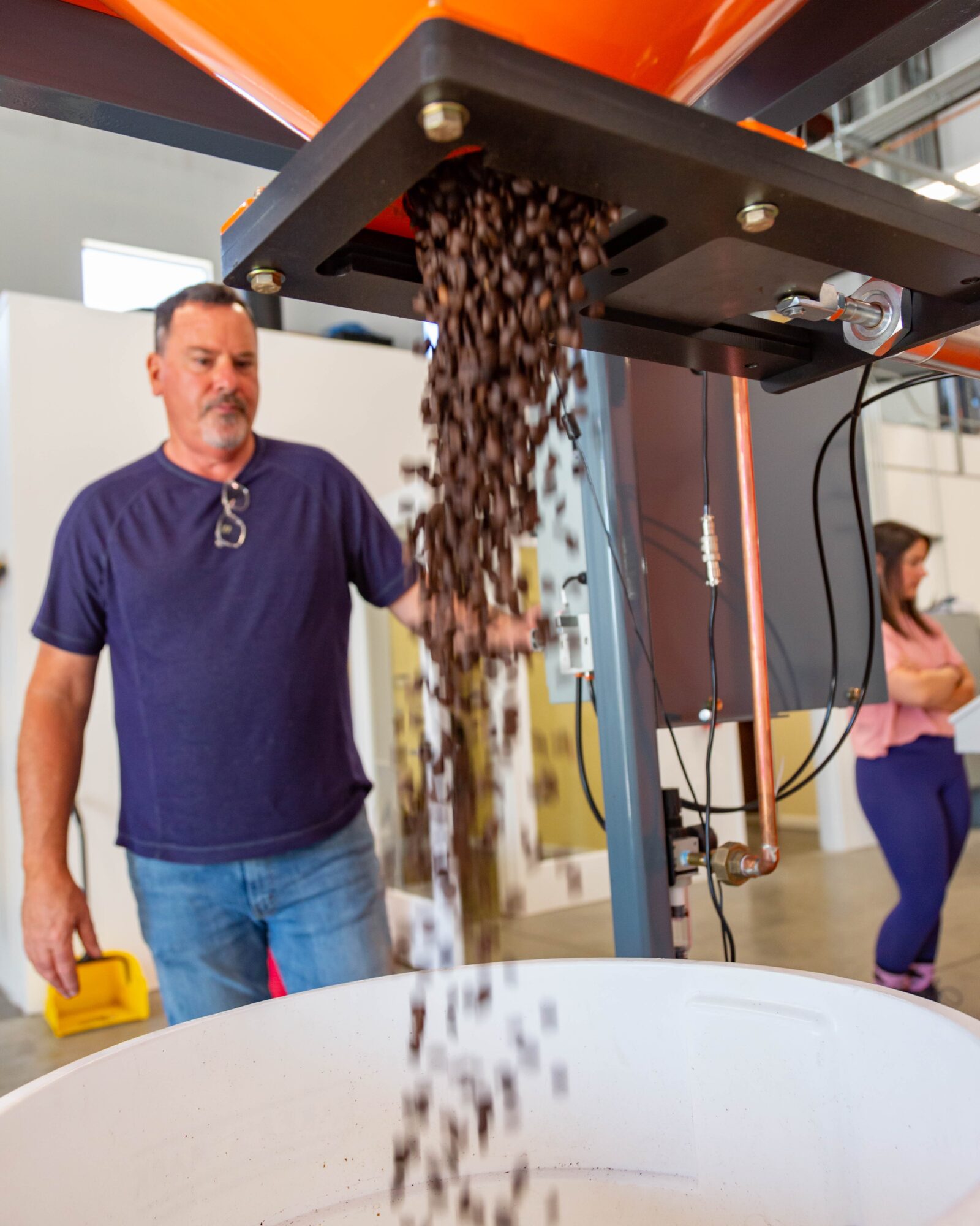 Man standing next to a machine pouring coffee beans into a container, with a woman in the background. Bright indoor setting.