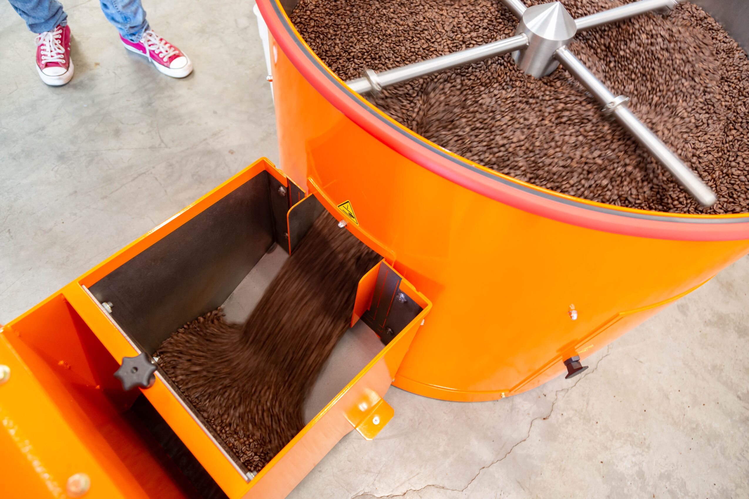 Close-up of an orange industrial coffee roaster with a metal drum and a chute filled with roasted coffee beans, with part of a person's shoes visible in the background.
