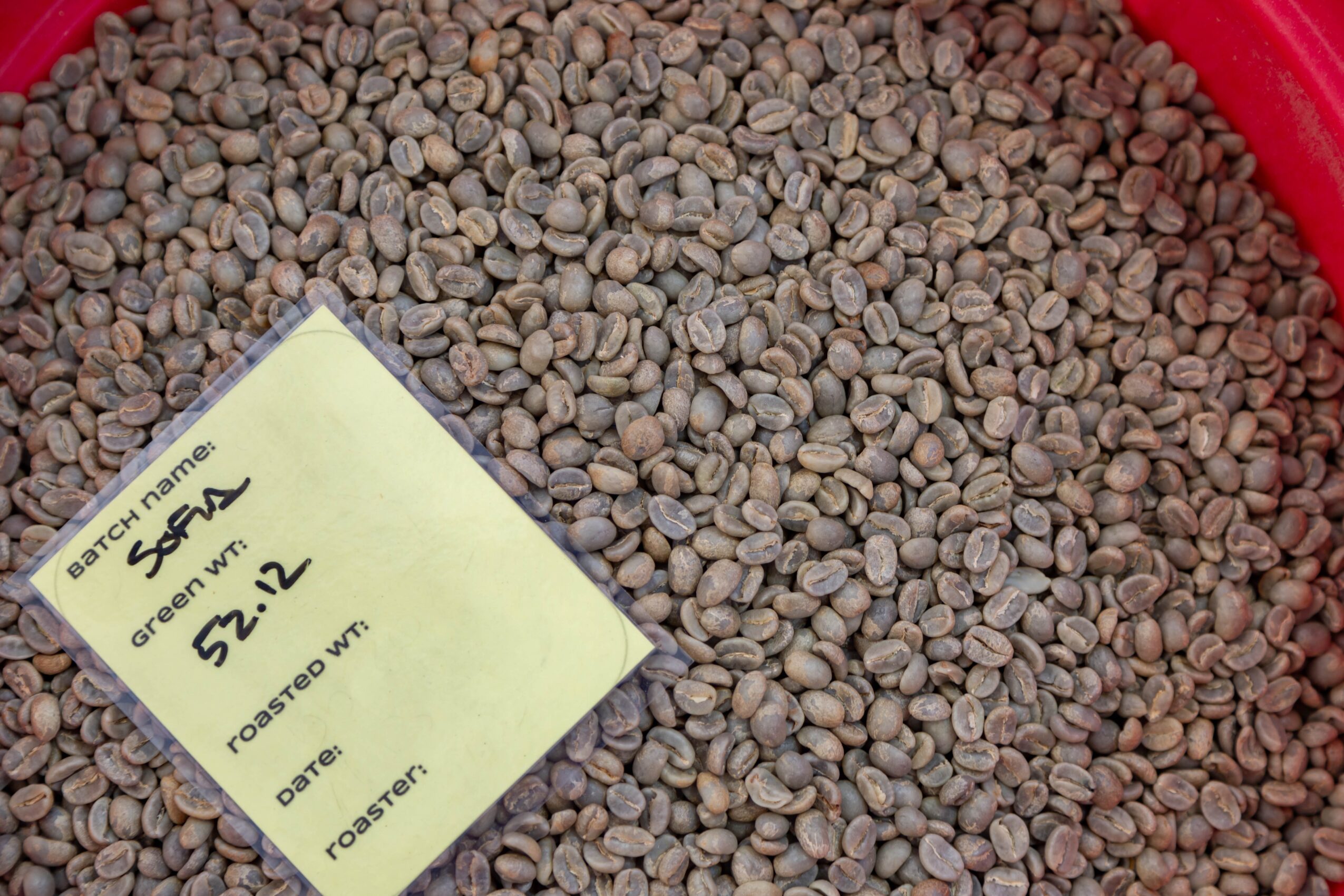 Close-up of green coffee beans with a yellow note labeled 'Brazilian beans, green, 52.2, roaster' on top.
