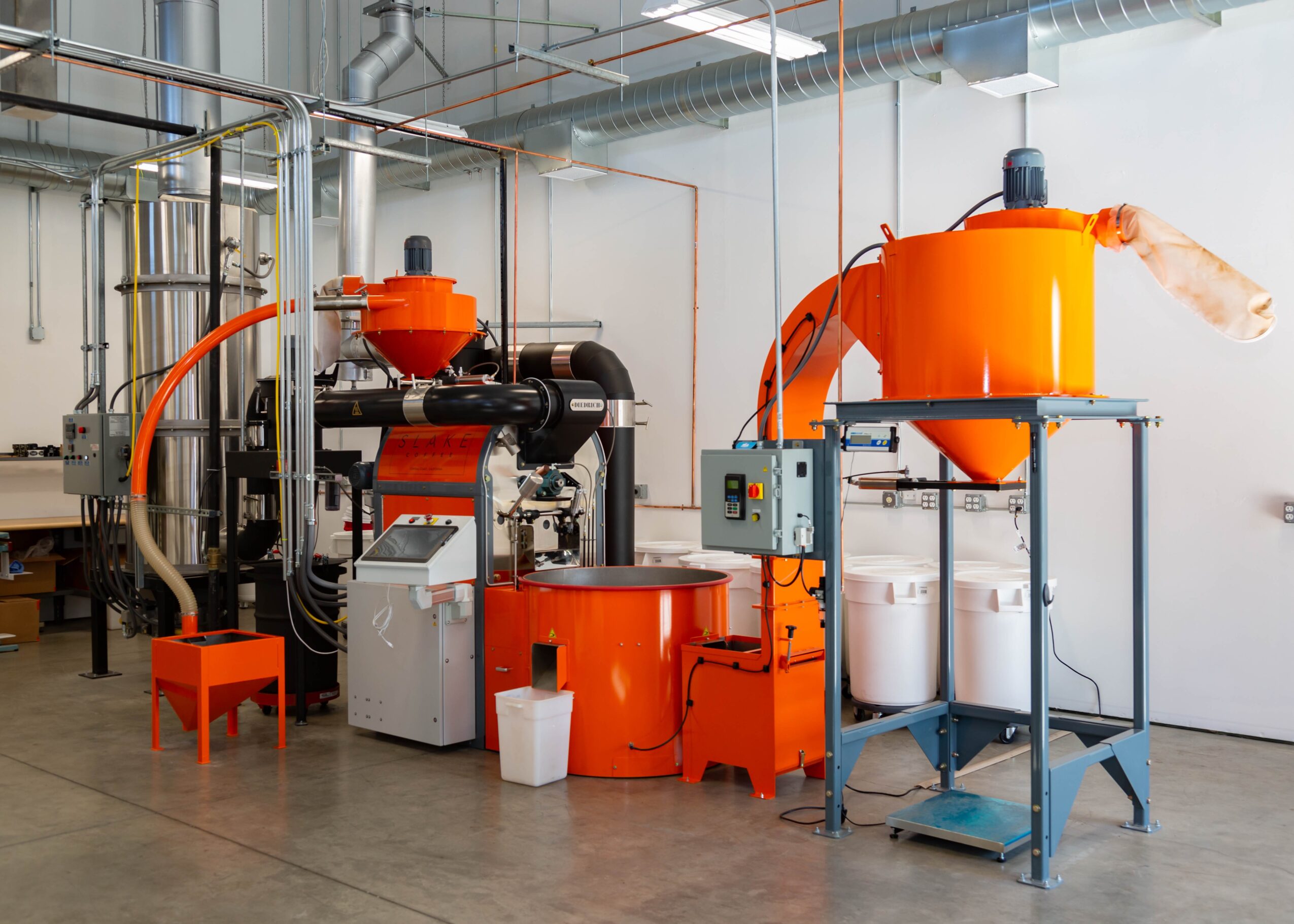 Industrial machinery with orange and black components in a factory setting, including a small orange chair and a white bucket.