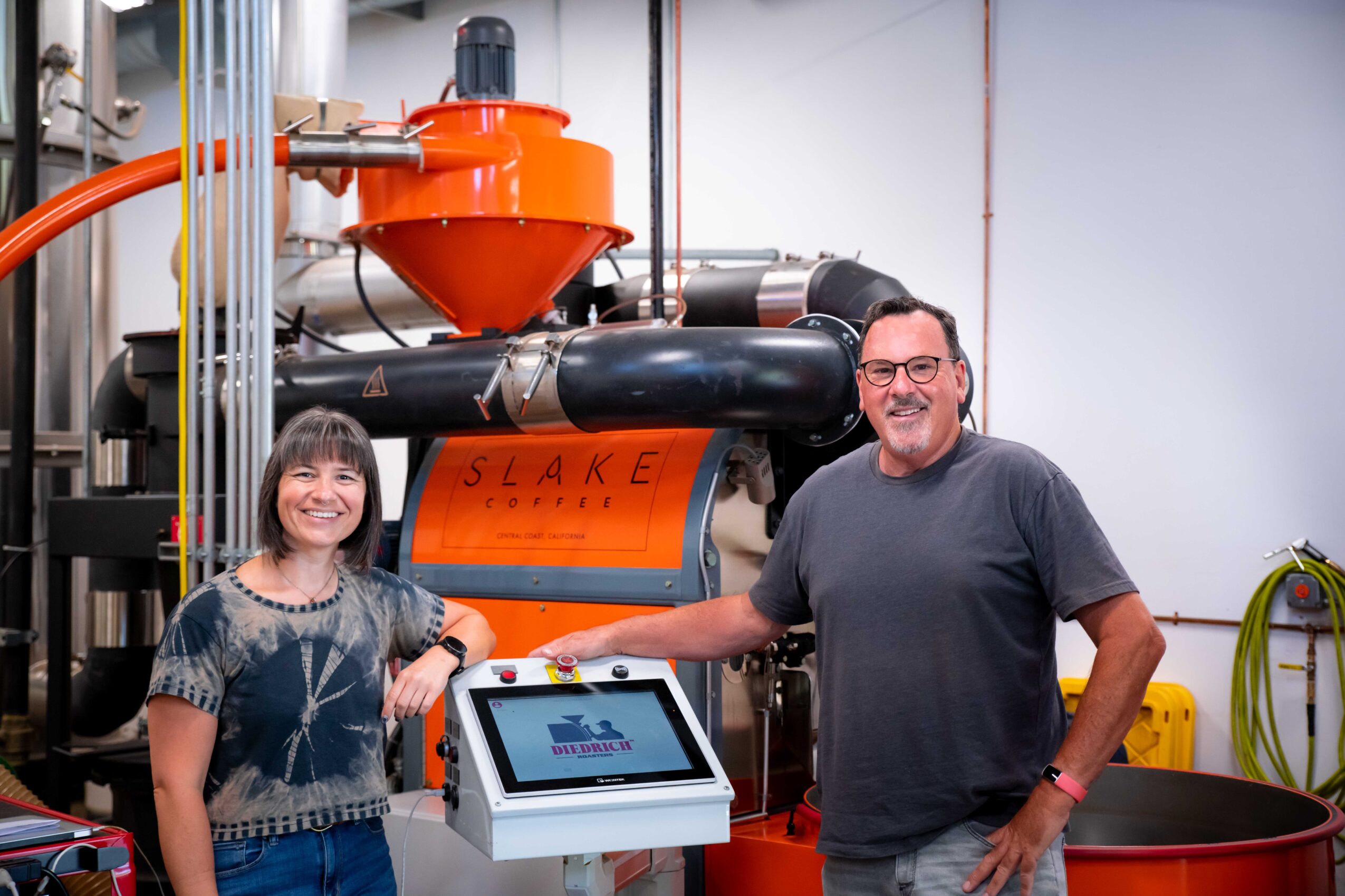 Two people stand next to industrial equipment, smiling, with a touchscreen device in front of them, in a factory setting.