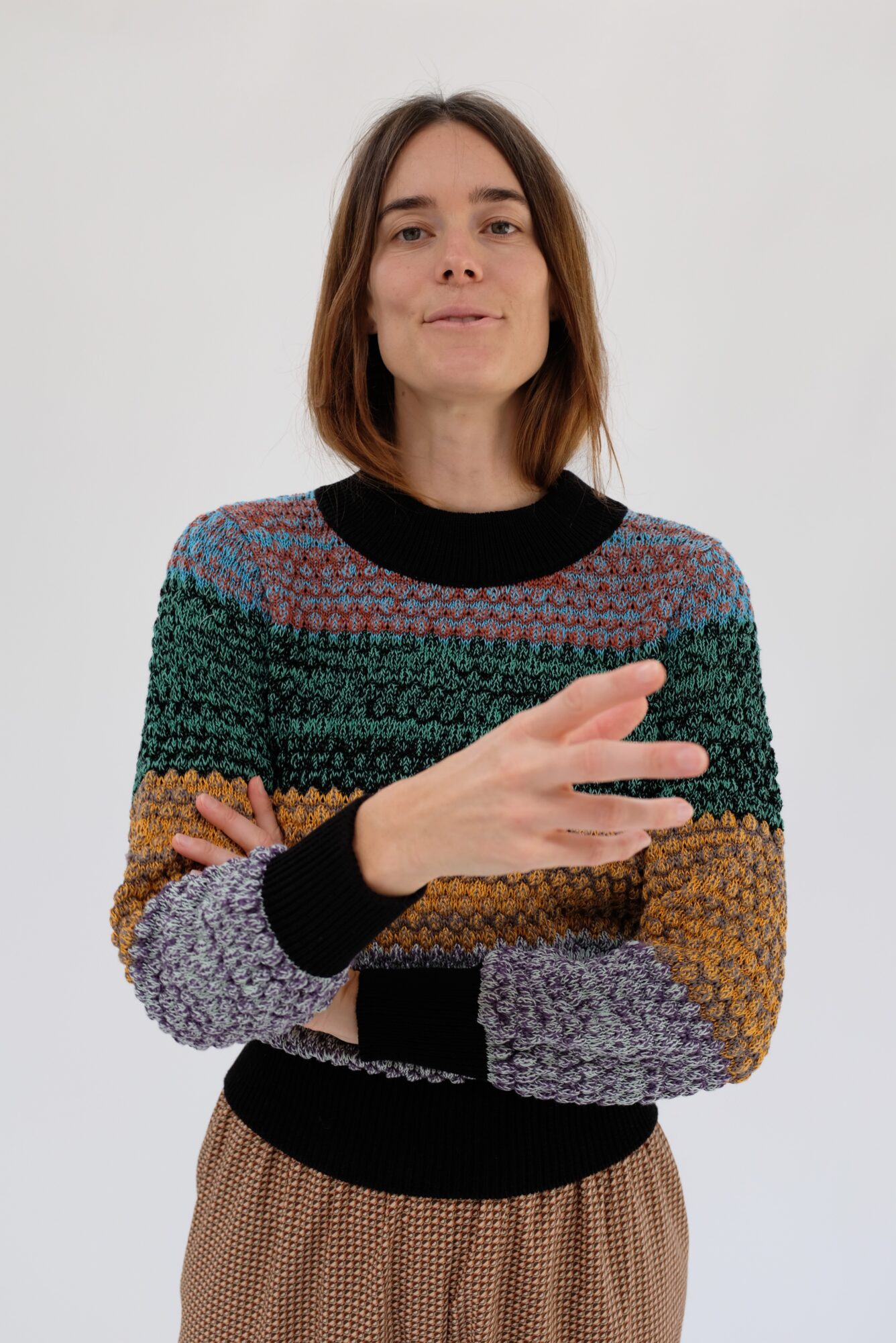 Woman with shoulder-length hair wearing a multicolored striped sweater, crossing arms, standing against a plain white background.